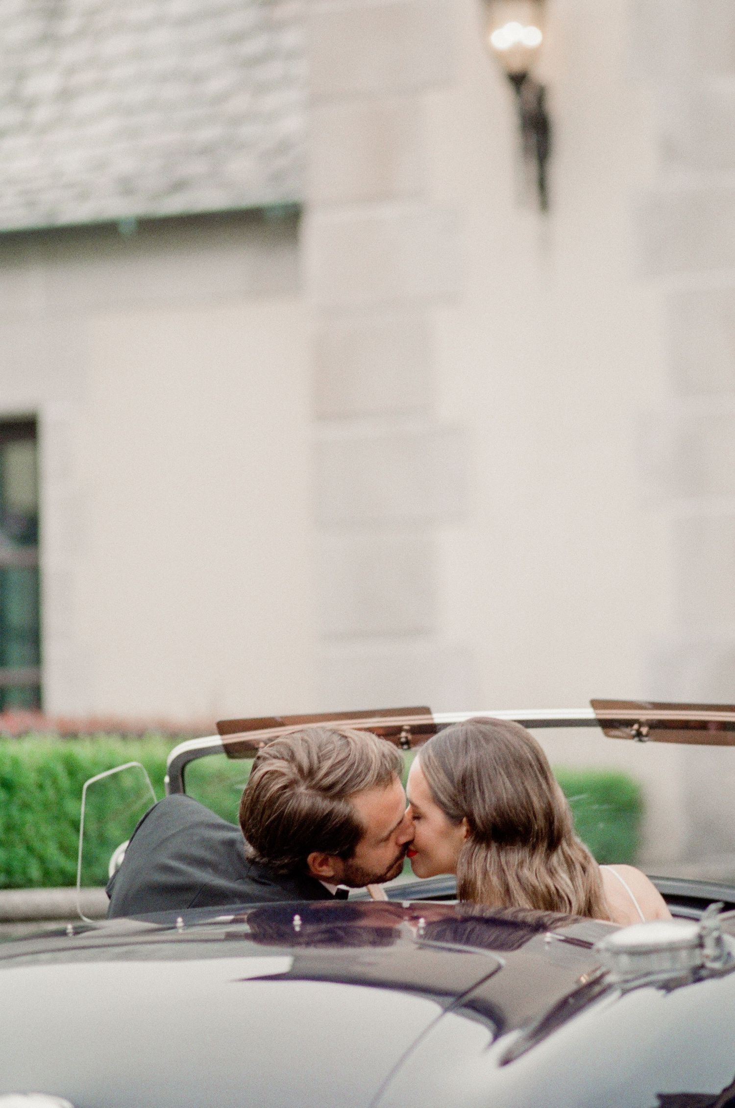A romantic couple shares a kiss while sitting in a vintage convertible car against a light stone building exterior.