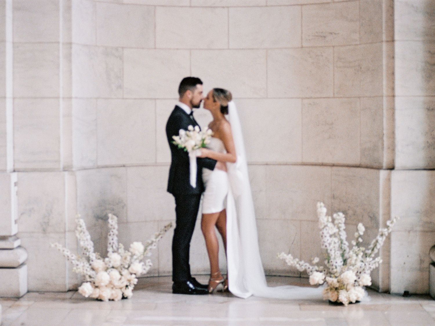 A bride and groom embrace in a minimalist stone alcove with white floral arrangements at their feet.