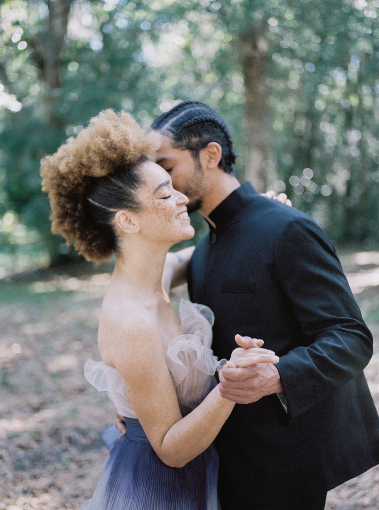 A couple shares an intimate dance moment outdoors in formal attire with natural lighting filtering through trees.