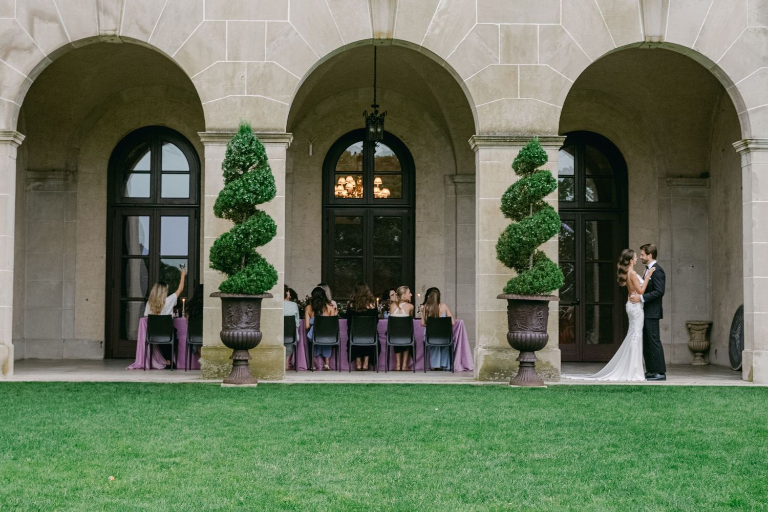 A couple shares a moment under elegant stone arches with wedding guests seated nearby and topiary plants decorating the entrance.
