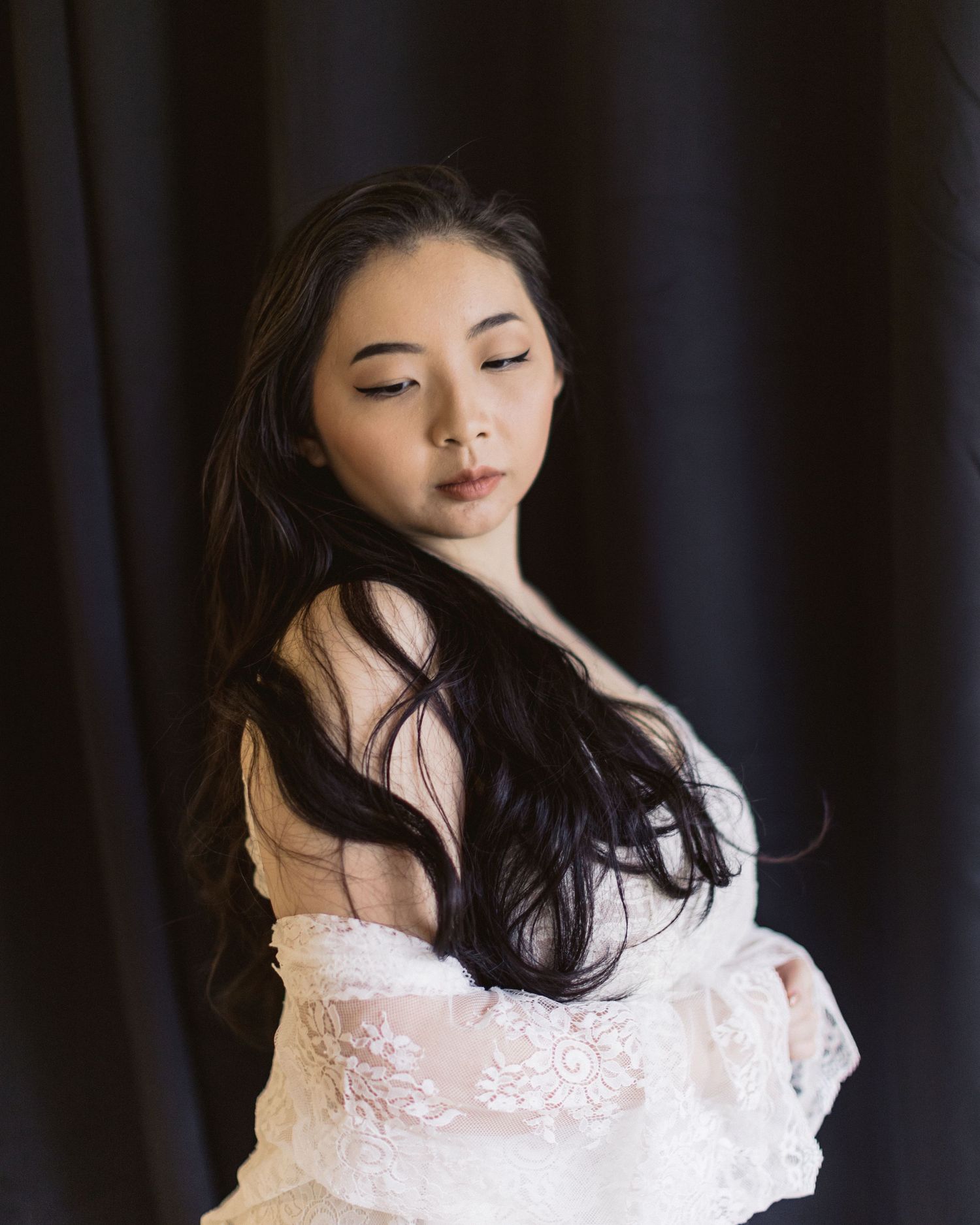 Moody portrait of young woman in white lace against dark backdrop with flowing black hair and natural makeup.