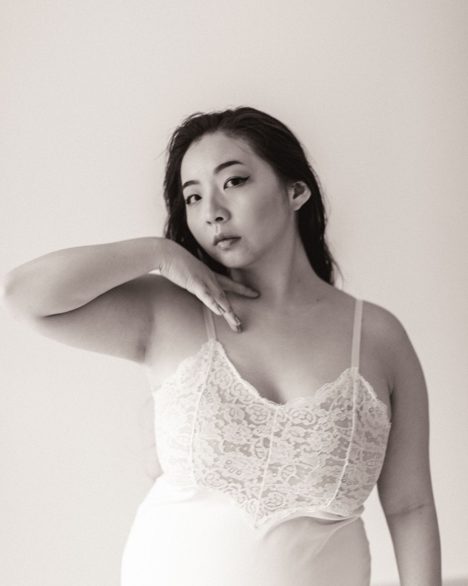 Black and white portrait photograph of a person in a white lace camisole posing with hand elegantly raised against neutral background.