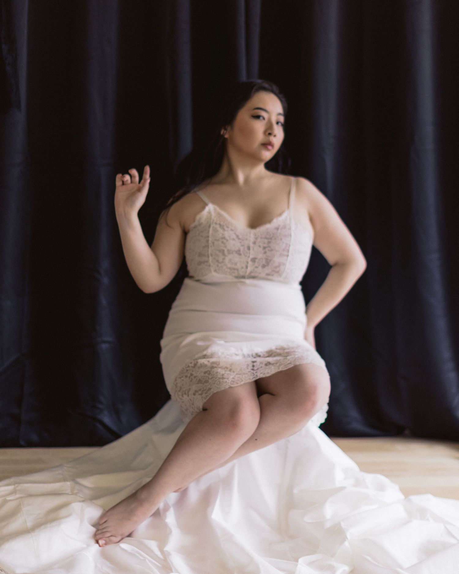 A curvy model poses elegantly in a white lace bridal lingerie and flowing skirt against a dark backdrop.