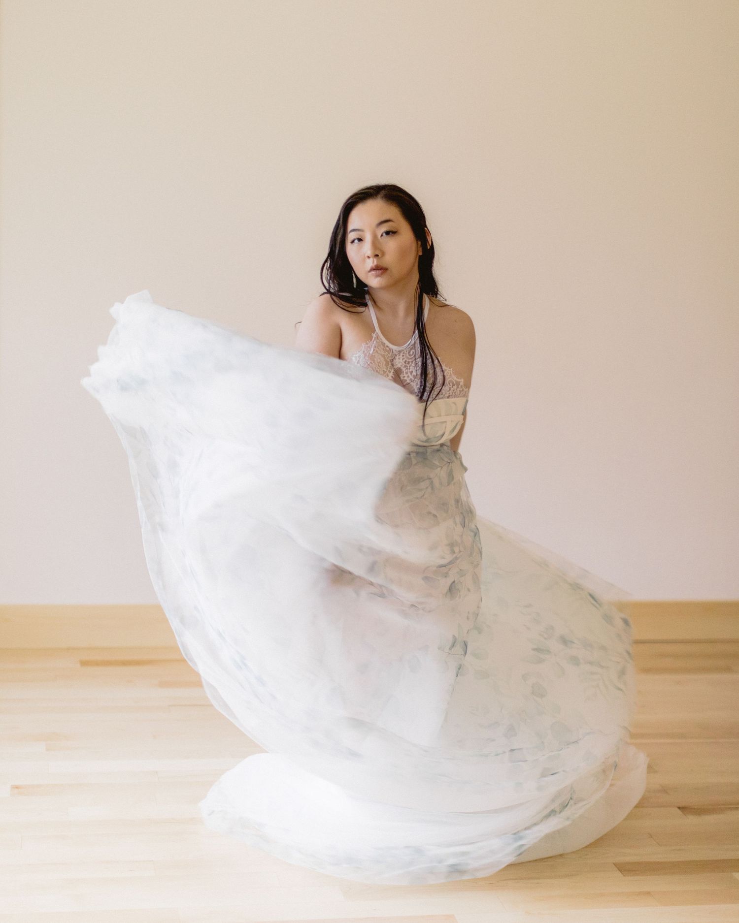 Elegant bridal portrait with flowing white tulle dress creating graceful movement on wooden floor against neutral background.