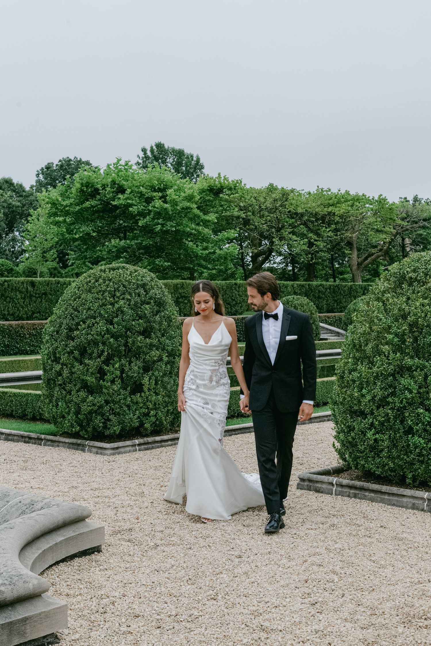 Newlyweds walk together on gravel path surrounded by manicured topiary bushes in elegant garden setting.