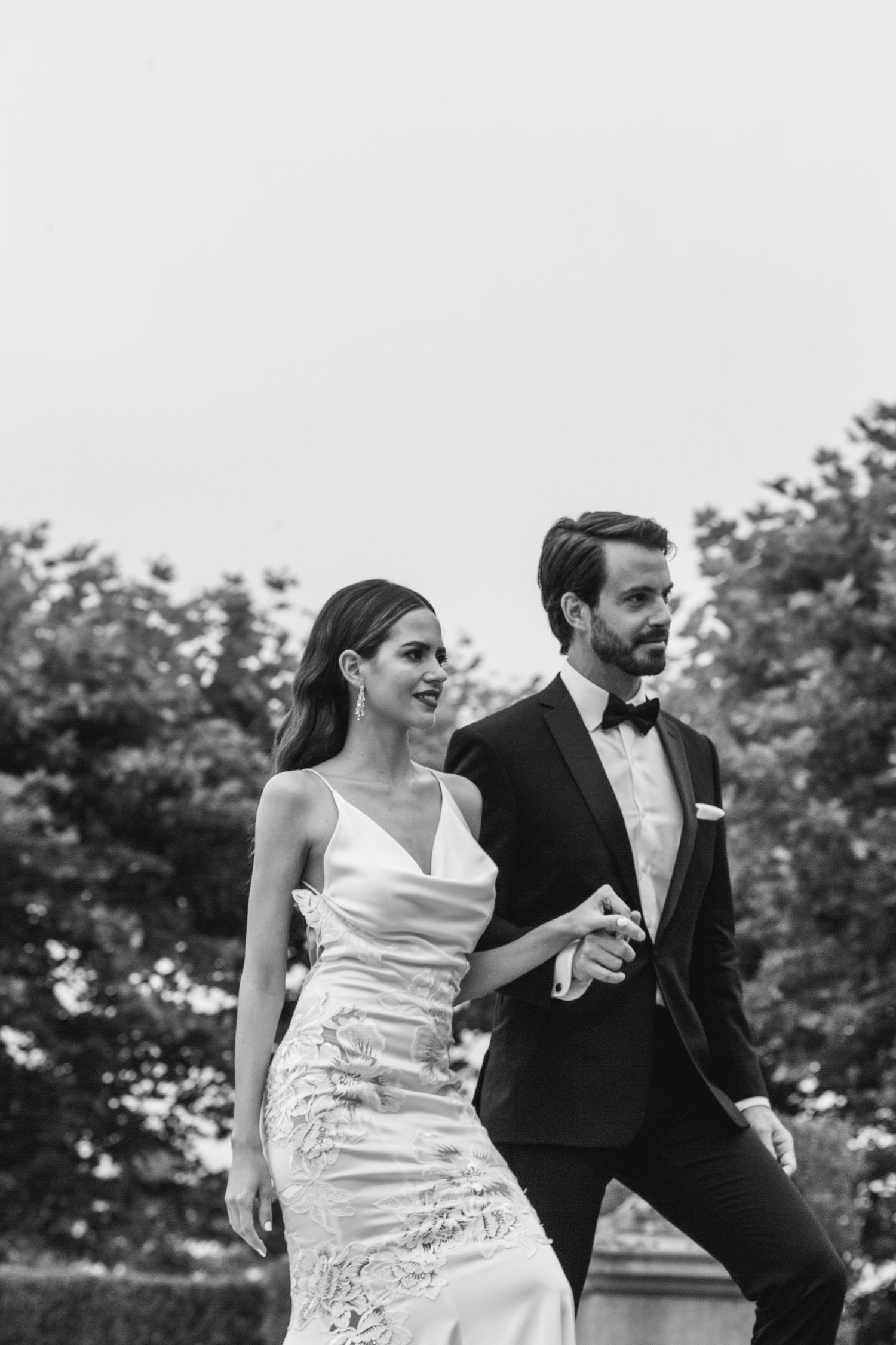 A black and white wedding photo shows a couple walking outdoors, with the bride in a fitted dress and groom in classic tuxedo.