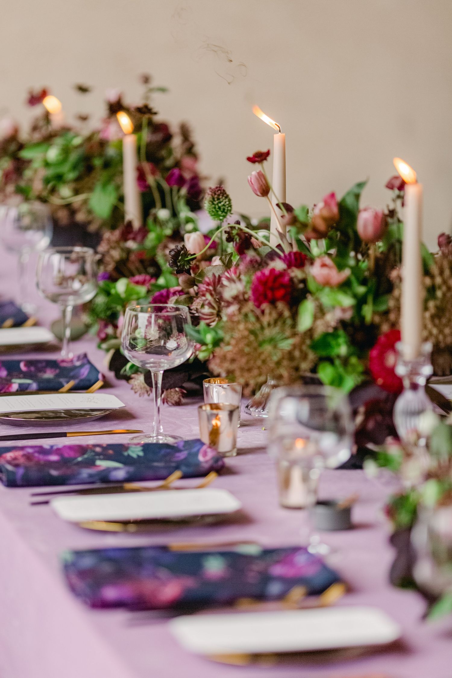 Elegant tablescape with lit taper candles, wine glasses, and colorful floral arrangements on a lilac tablecloth.