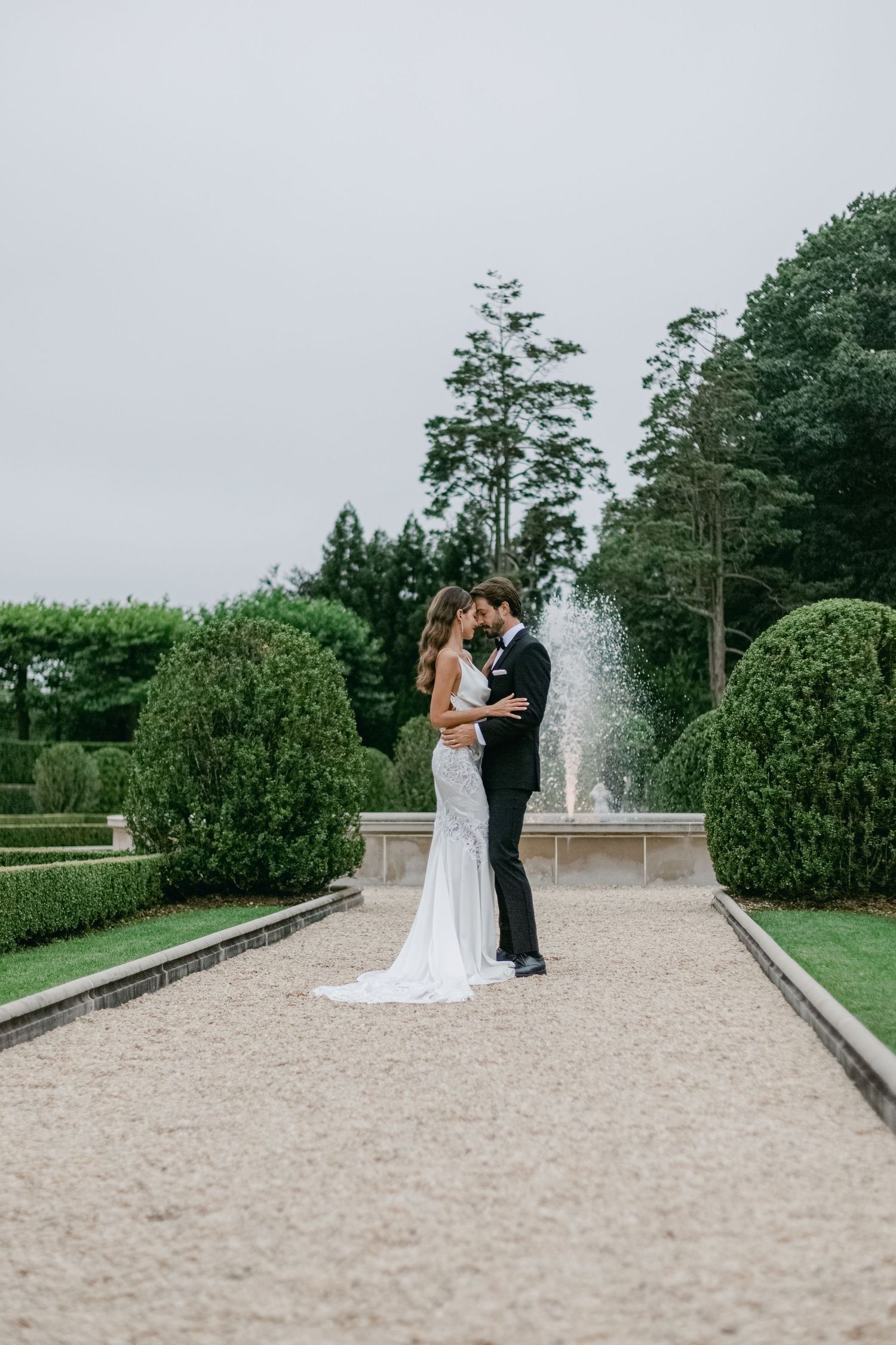 A romantic wedding couple embraces on a gravel path surrounded by manicured gardens and a fountain in the background.