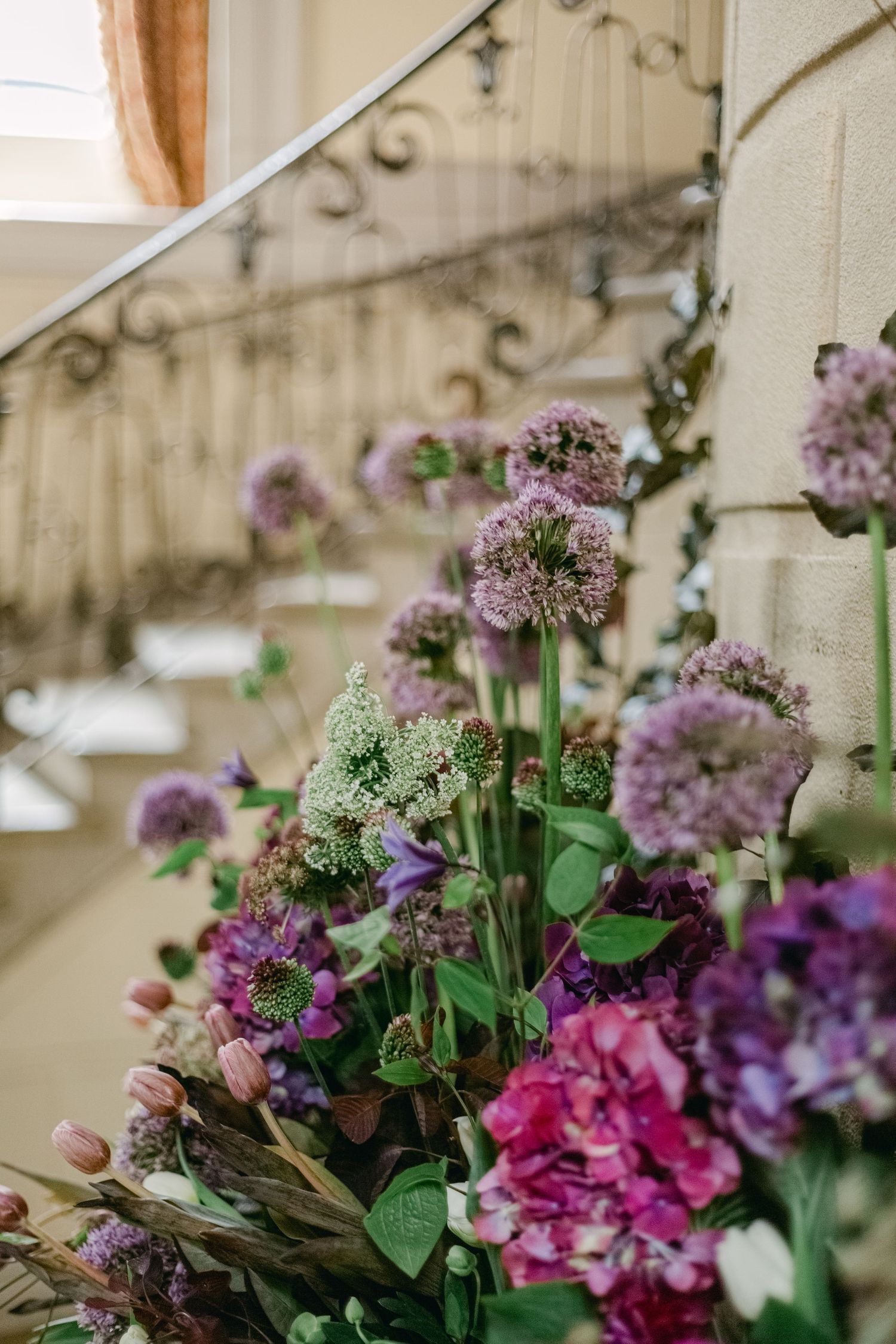 Beautiful floral arrangement with purple alliums, pink hydrangeas and greenery against a light interior wall and banister.
