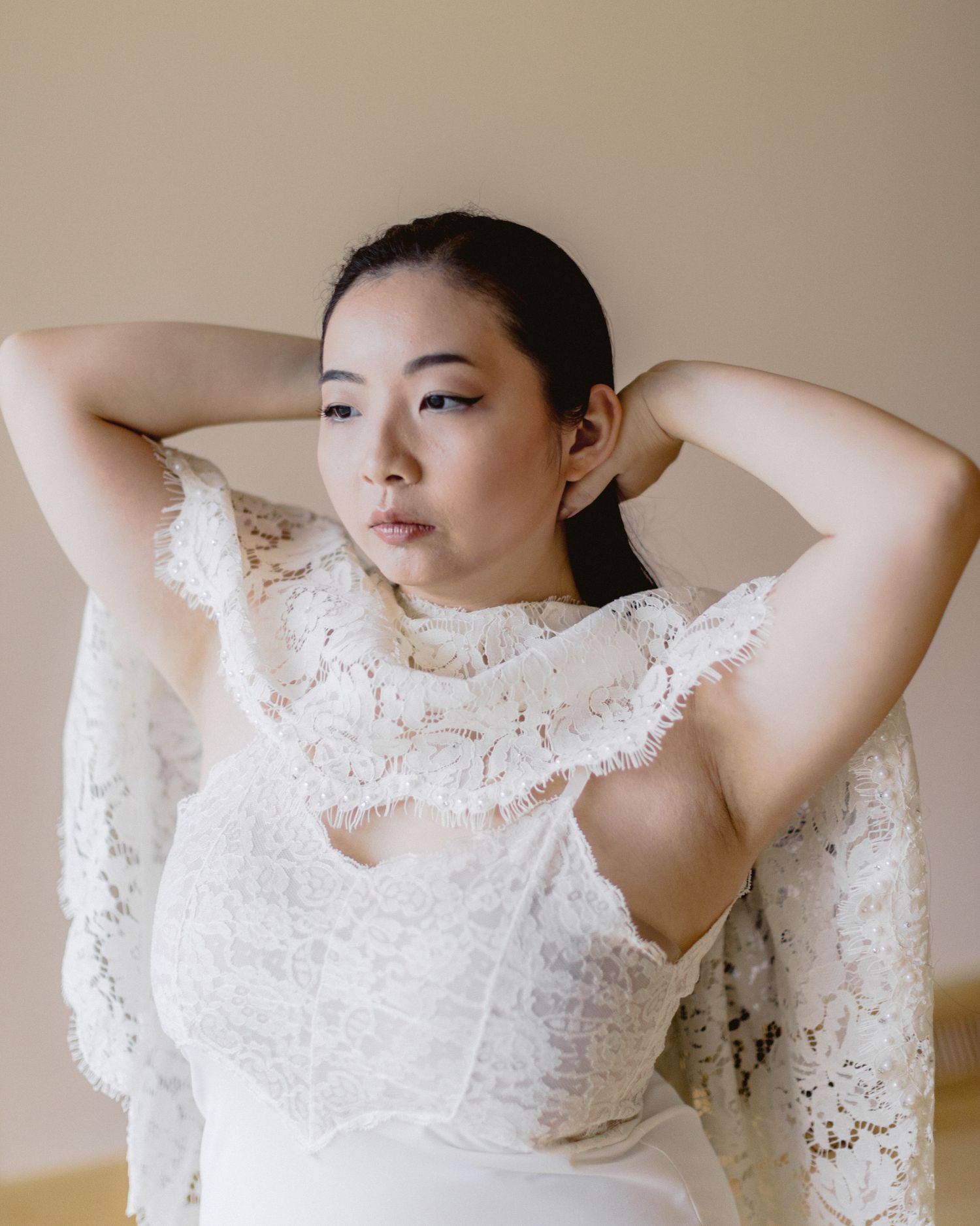 A person wearing an elegant white lace bridal gown with intricate details poses against a neutral background.