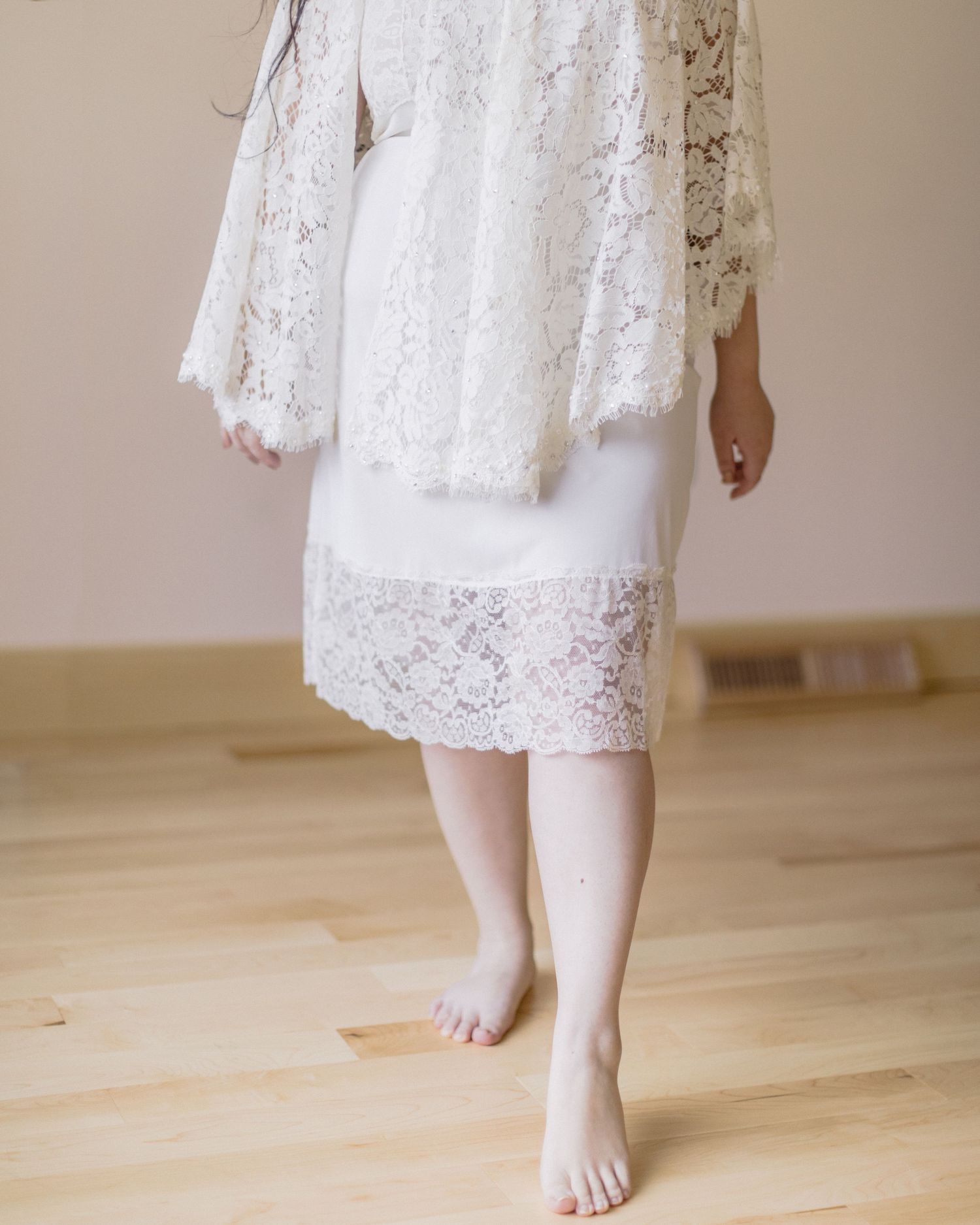 Bare feet and white lace dress hem standing on light hardwood flooring with a delicate flowing skirt.