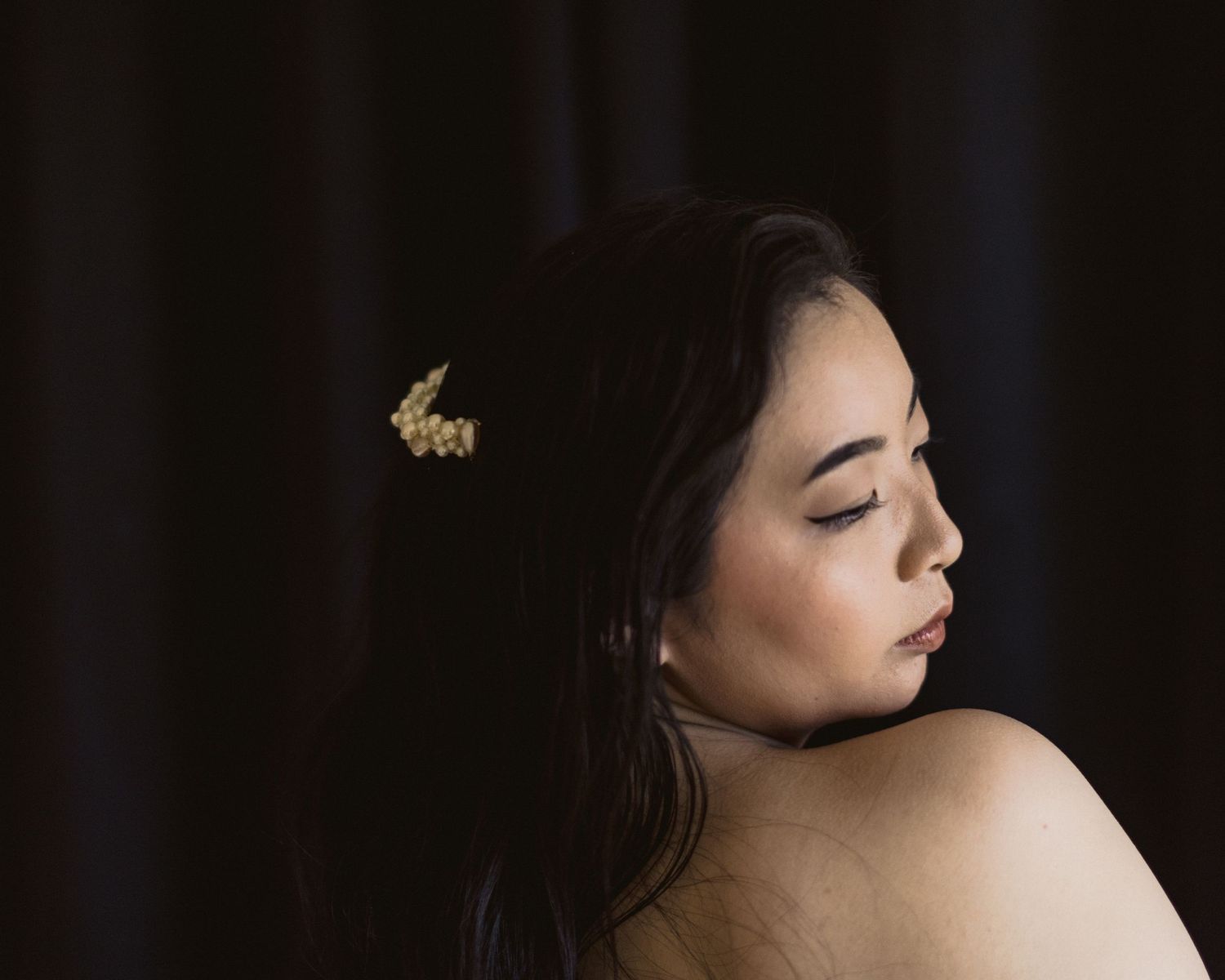 Side profile portrait in moody lighting showing bare shoulders and long dark hair with a butterfly hair accessory.