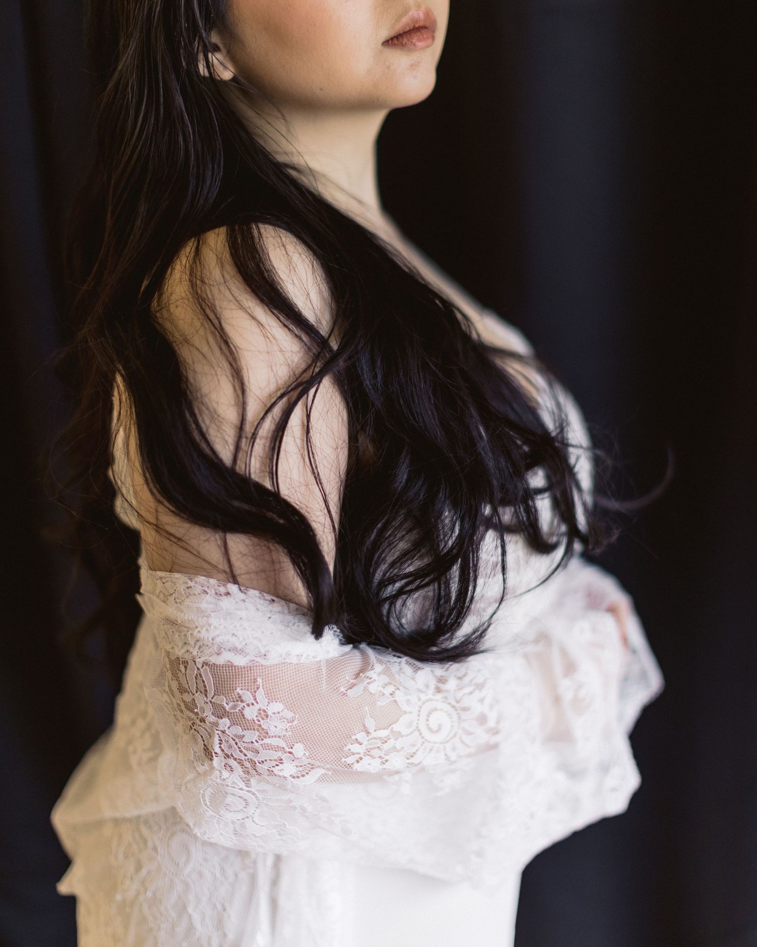 Dark-haired figure in white lace formal wear photographed from the side in dramatic lighting.
