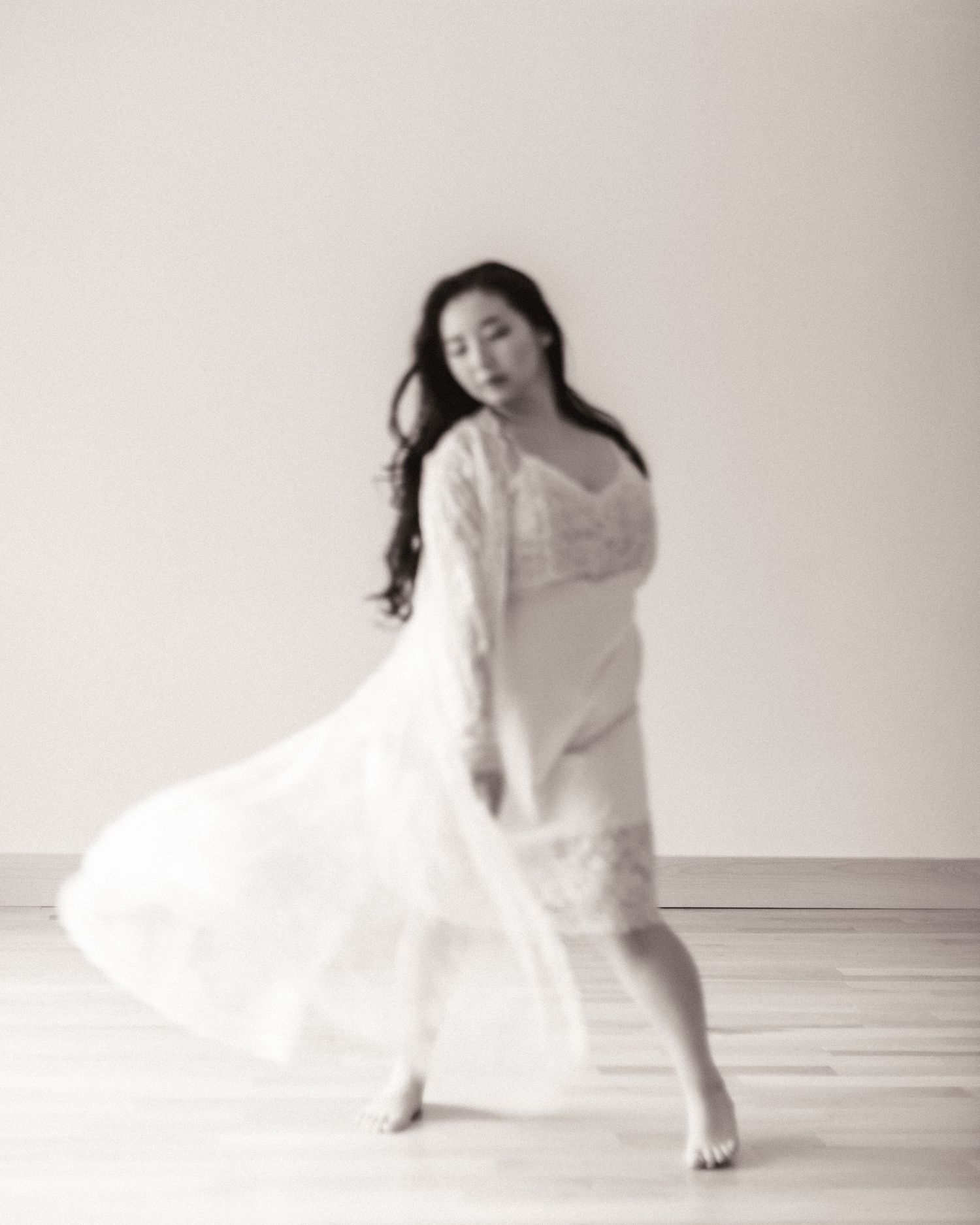 A graceful dancer in white flowing dress performs an expressive movement in a minimalist studio setting.