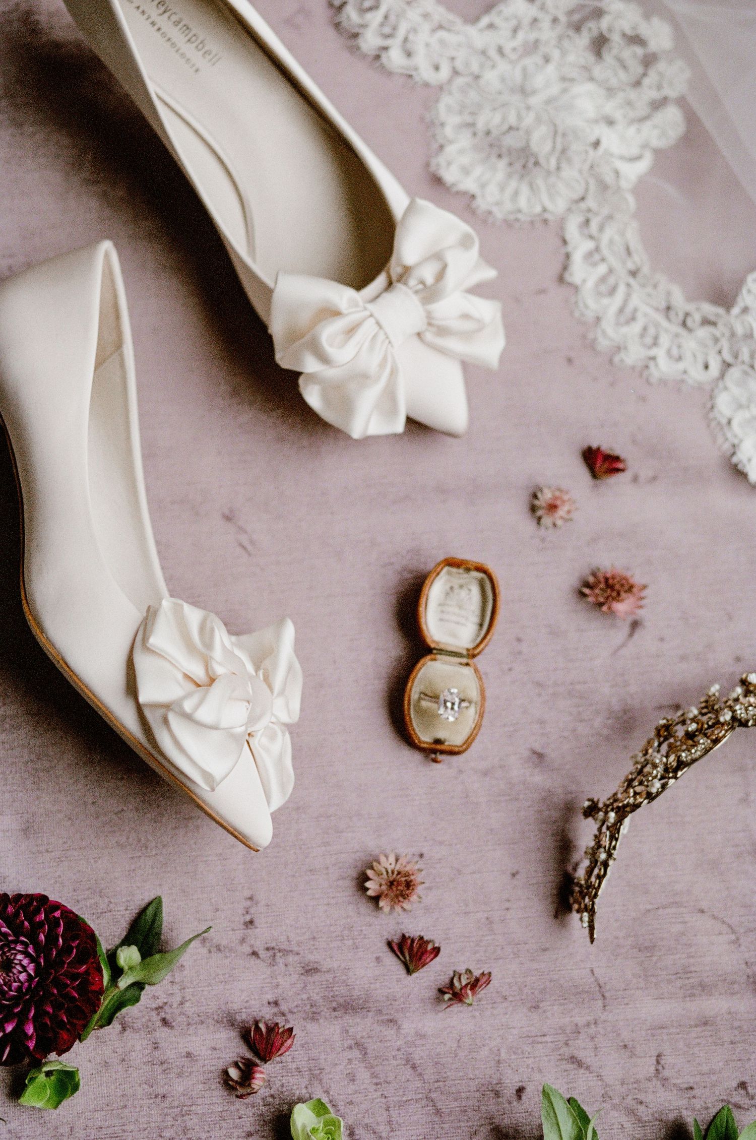 White bridal shoes with bows displayed alongside a vintage ring box and dried flowers on a pink textured surface.