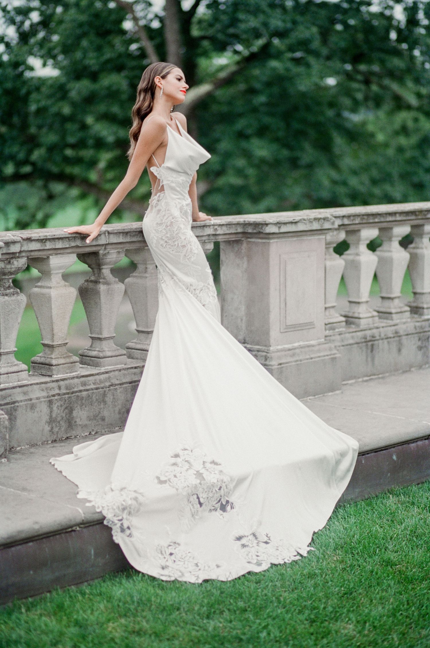Elegant mermaid wedding dress with lace details flows gracefully against stone balustrade in outdoor garden setting.