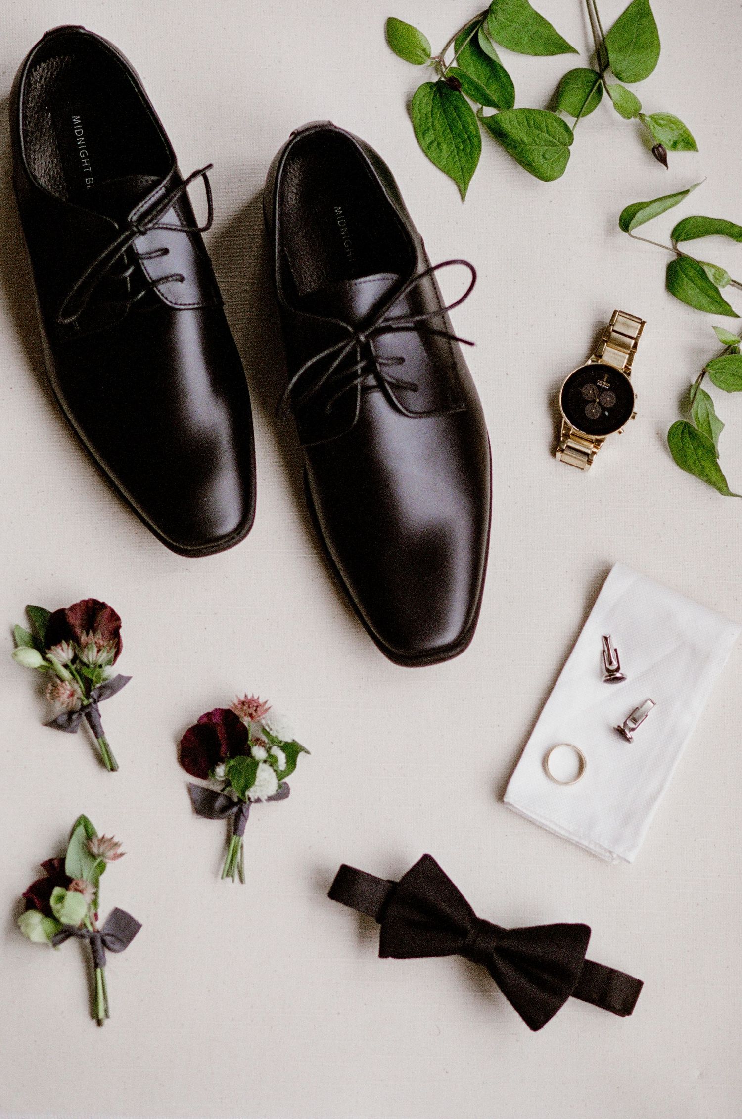 Elegant black leather dress shoes arranged with boutonniere flowers, bow tie, green leaves, watch and cufflinks on white surface.