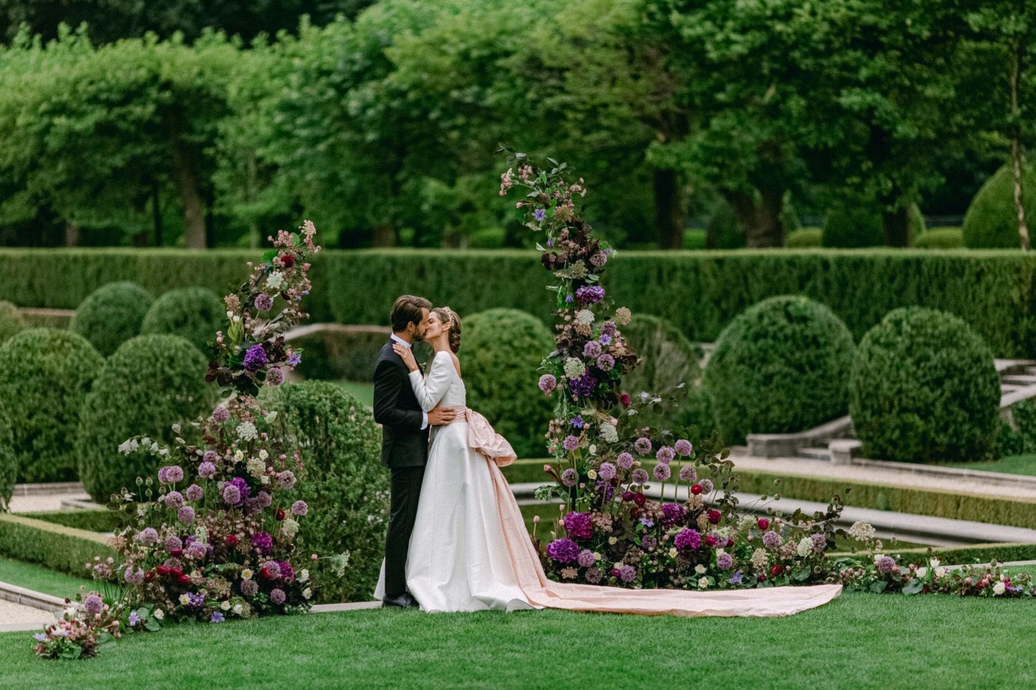 Elegantly dressed couple embraces in a manicured garden with floral arches and trimmed hedges on a sunny day.