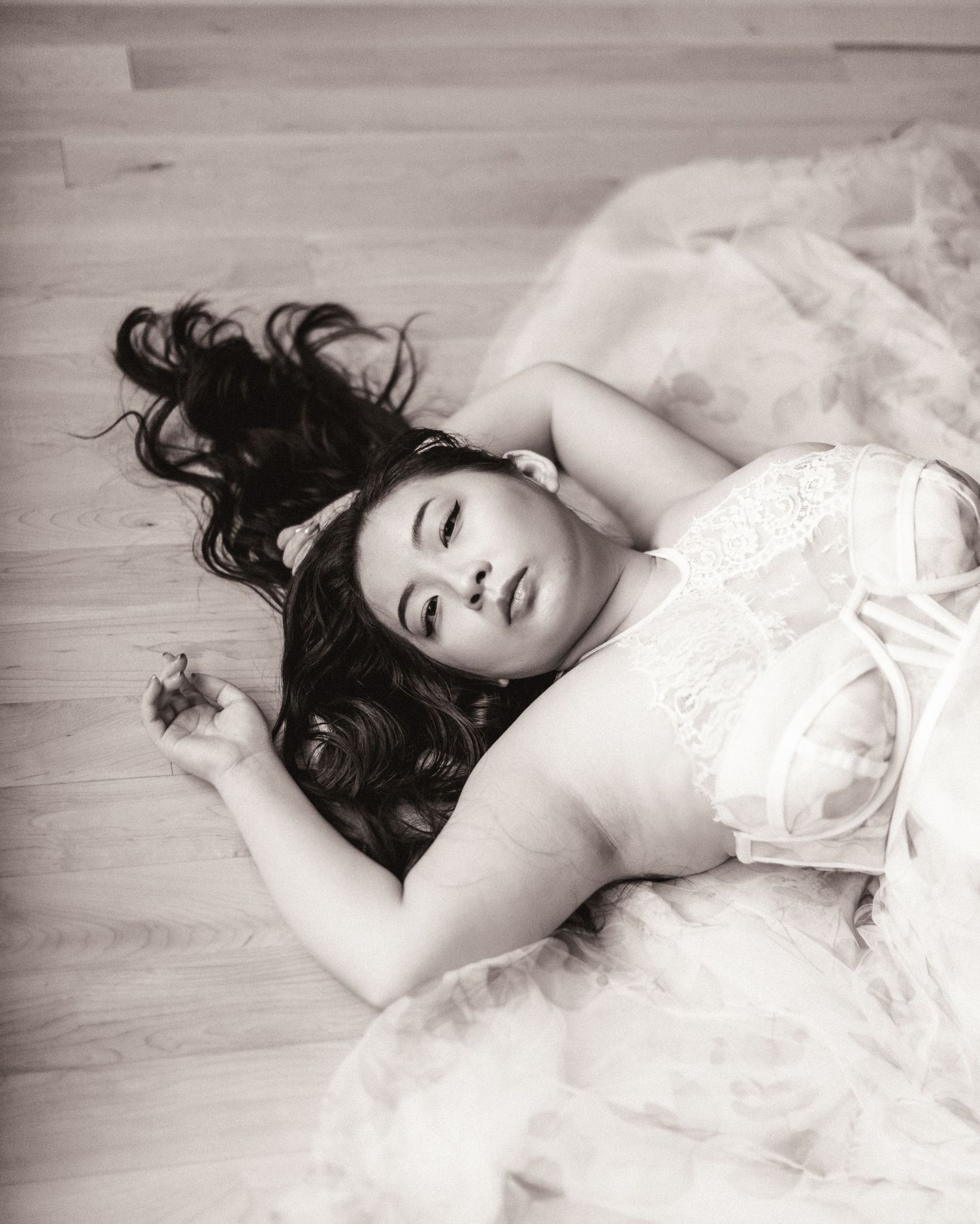 A black and white portrait shows someone lying on a wooden floor in a flowing white dress with long dark hair splayed out.
