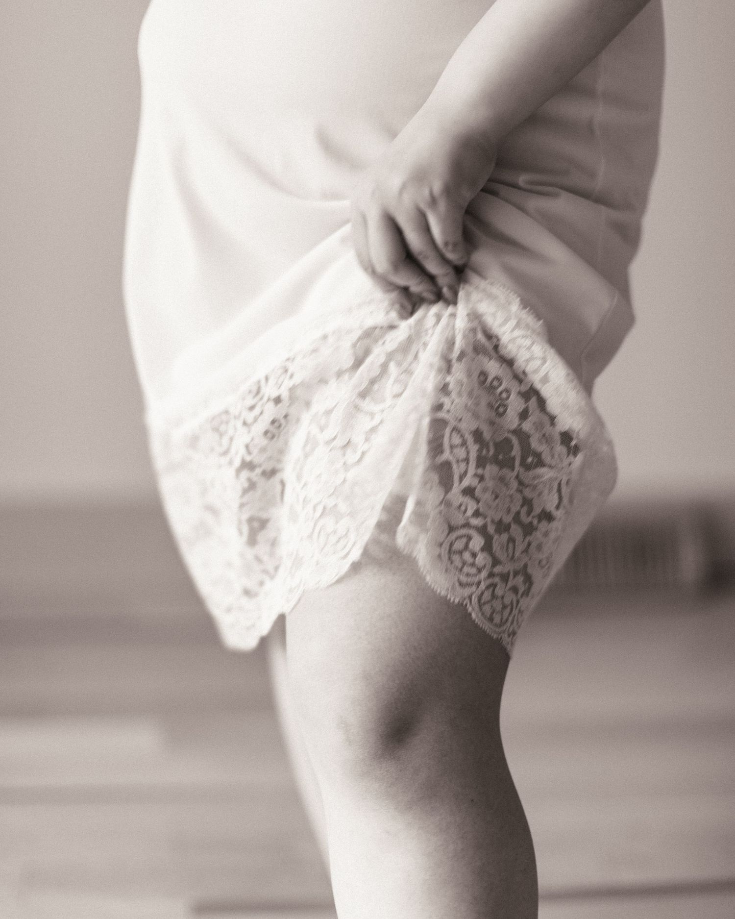 Close-up black and white photo of delicate lace details along the hem of a white dress, showing feminine elegance.