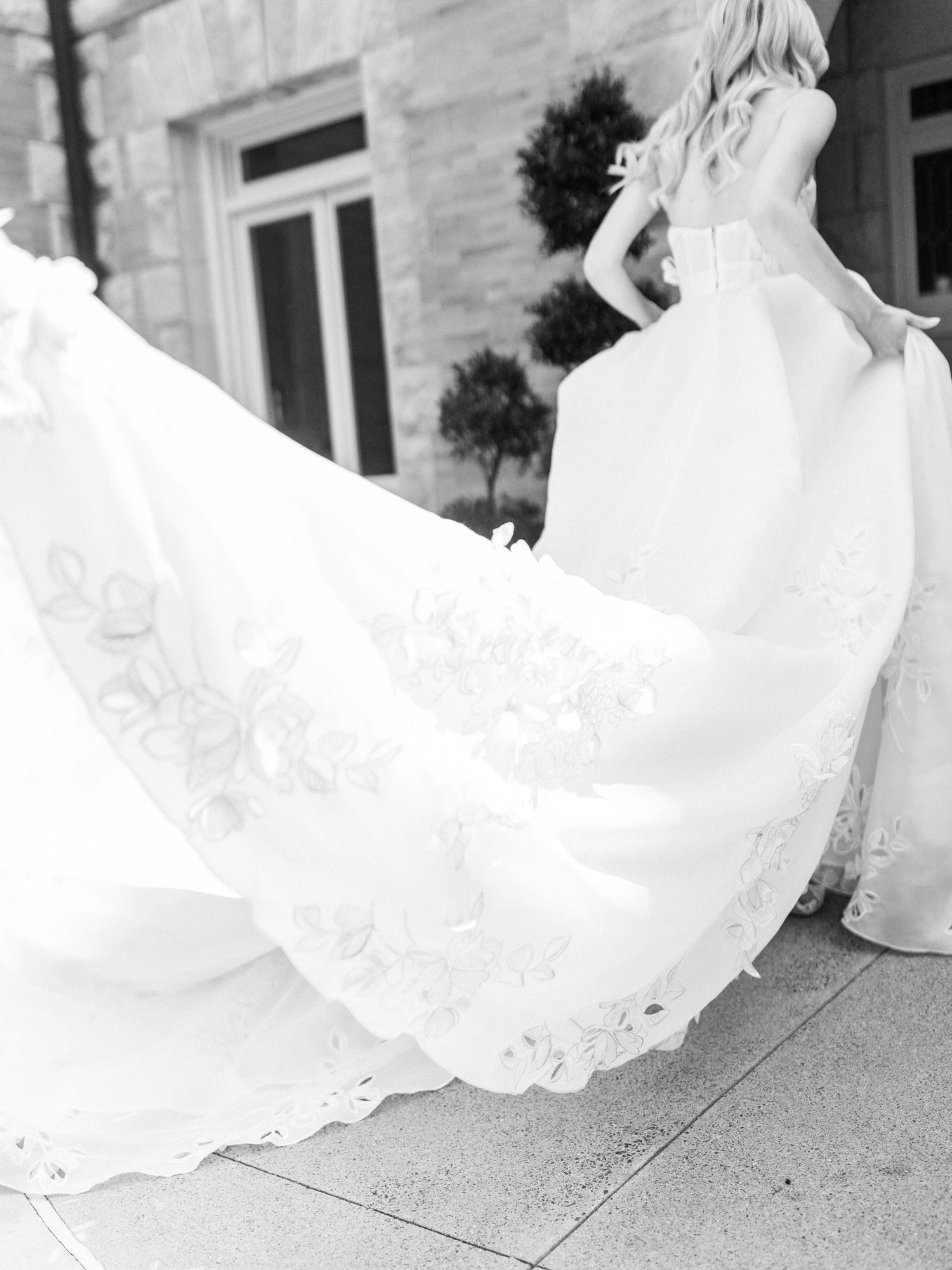Black and white photograph showing the flowing train of an ornate lace wedding dress against a stone exterior.