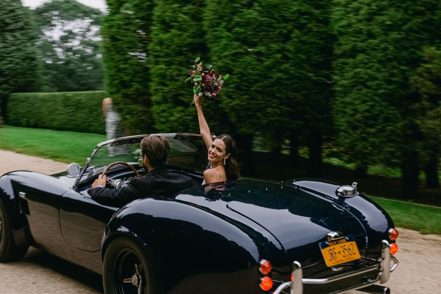 A black vintage convertible sports car parked on a tree-lined driveway with hands raised in celebration.