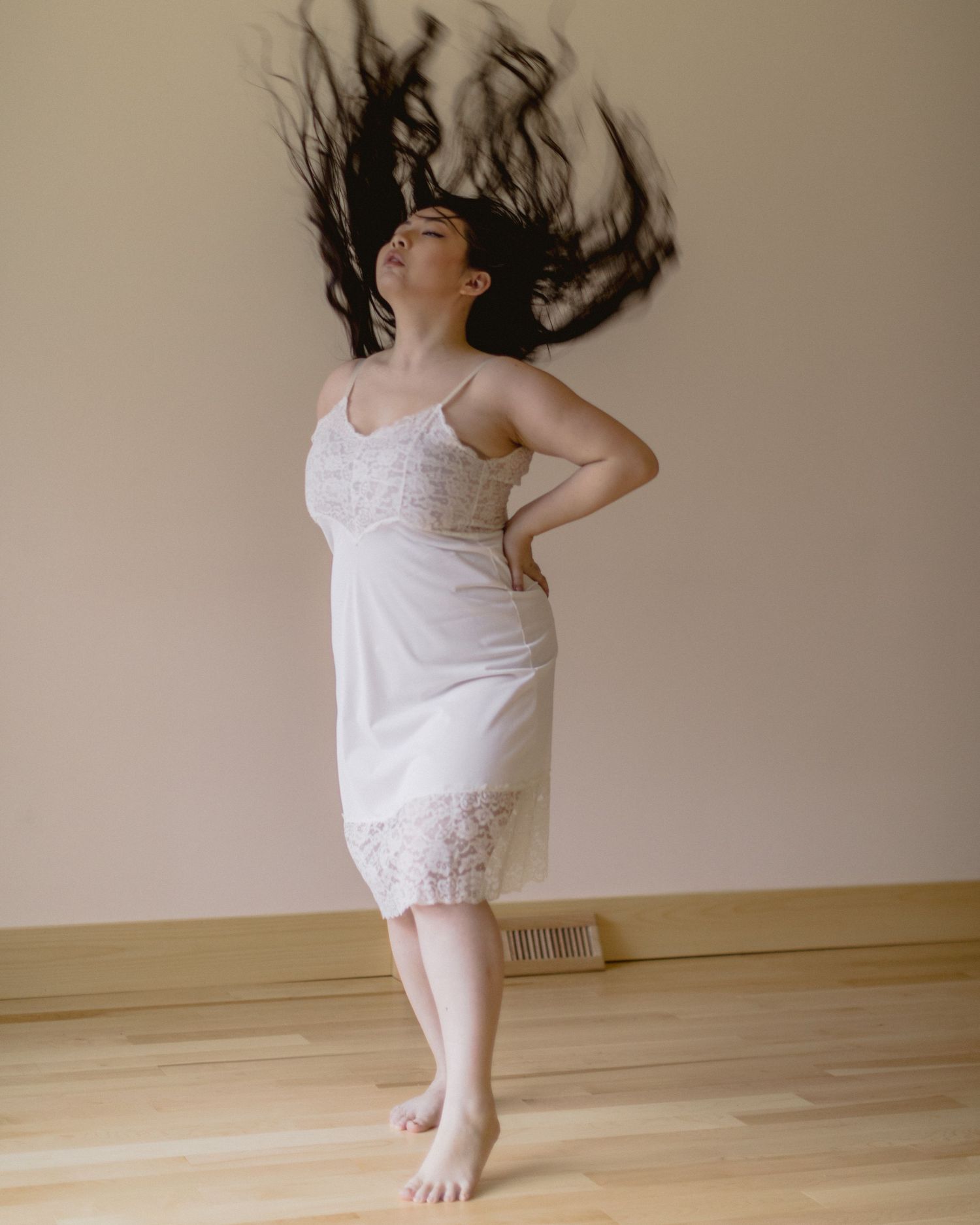 Person in white lace slip dress with flowing dark hair jumping and dancing on hardwood floor against beige wall.