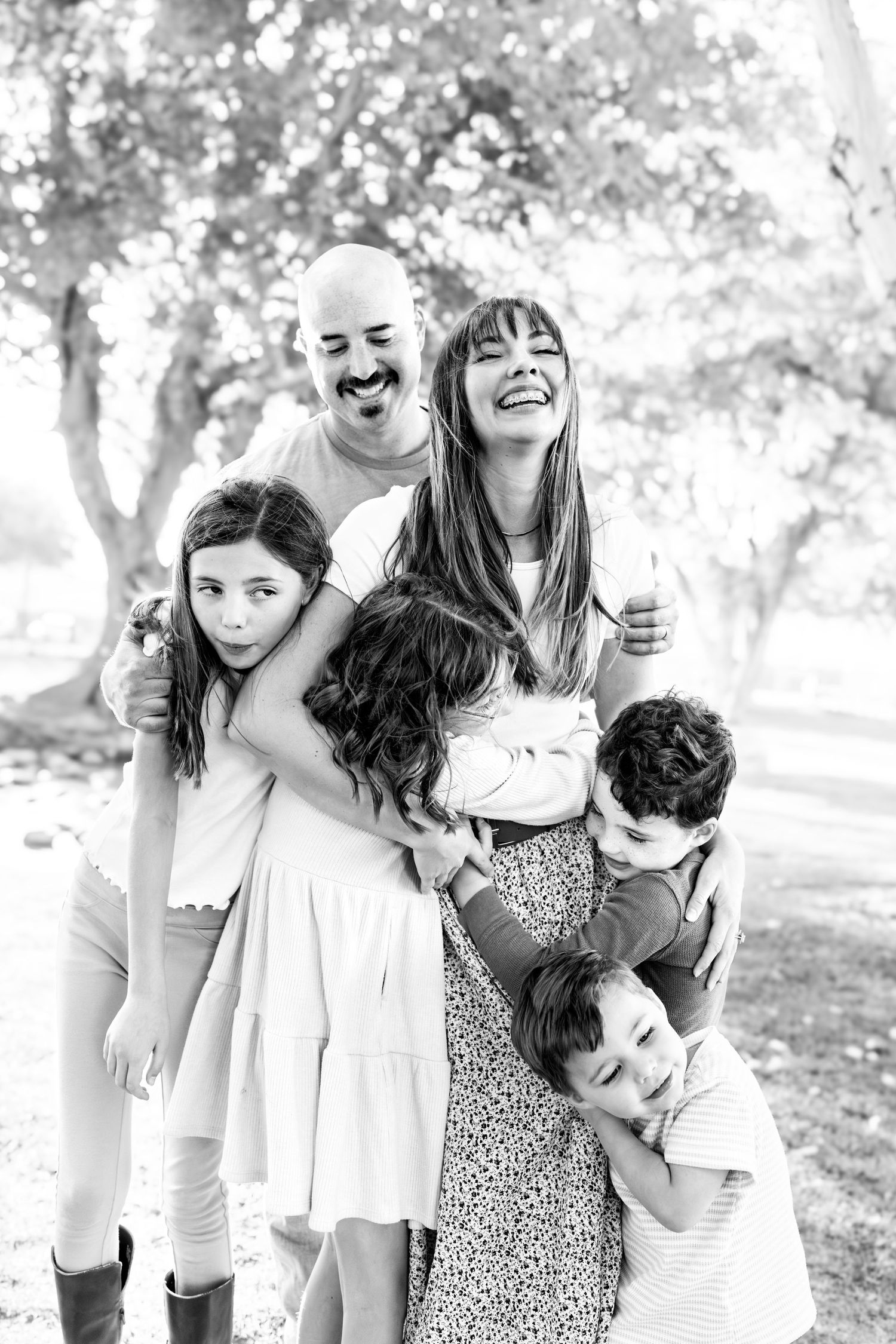 black and white image of a mother laughing with her head back as her 4 children embrace her in a huge hug with dad hugging her from behind