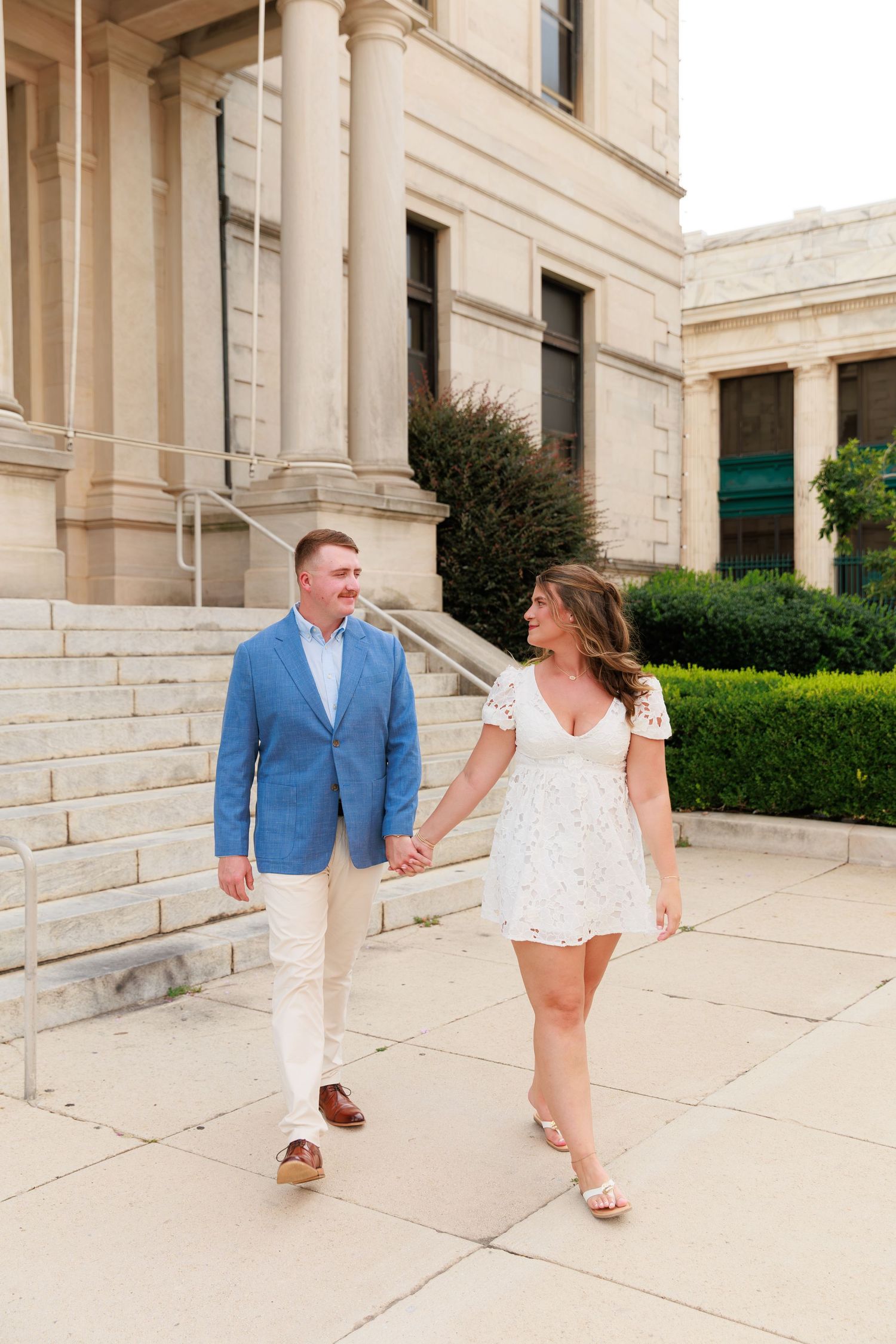 Smiling couple holds hands walking outside a classical stone building, man in blue blazer and woman in white dress.