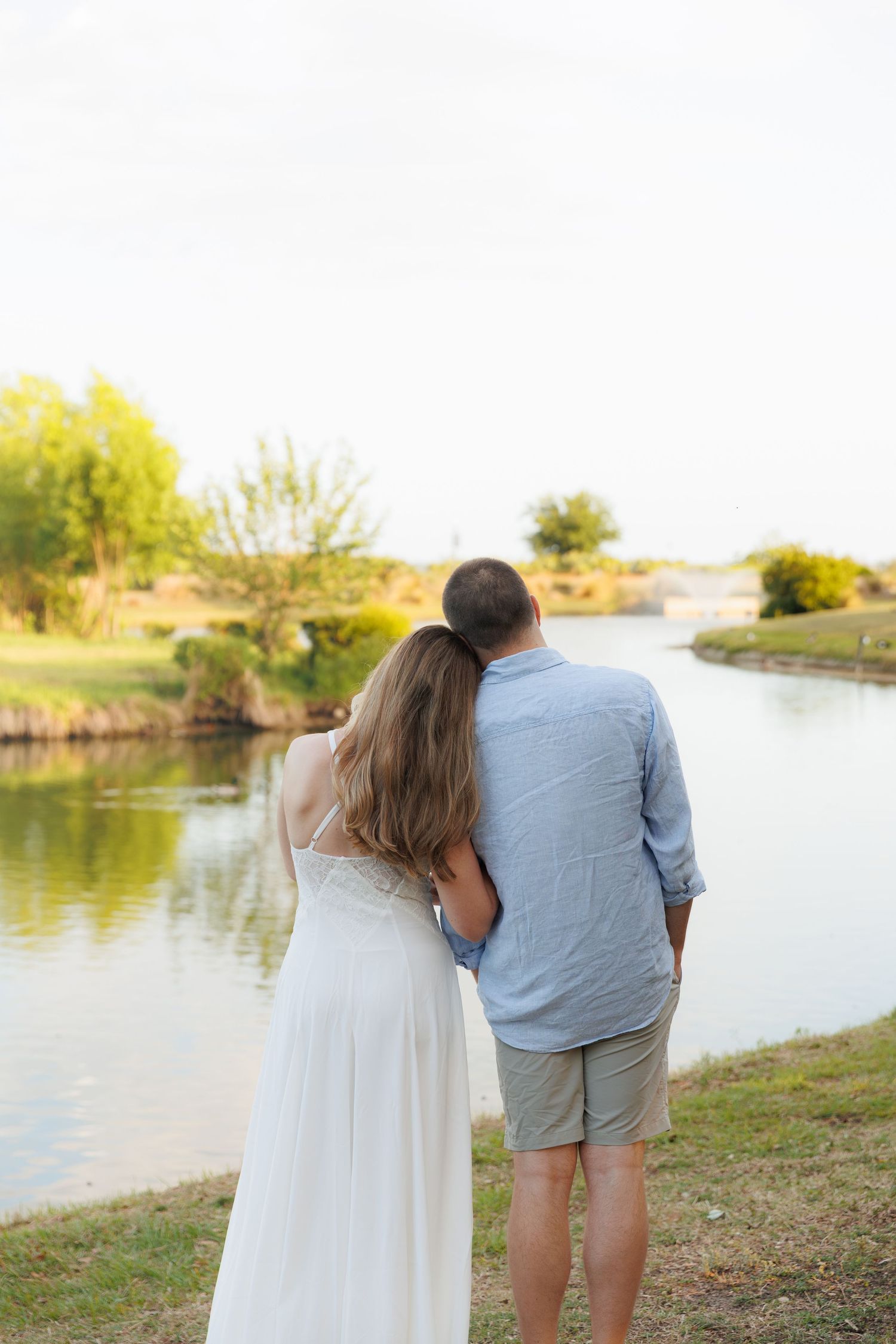 Couple standing arm in arm with backs to camera, gazing at a peaceful pond surrounded by green trees at sunset.