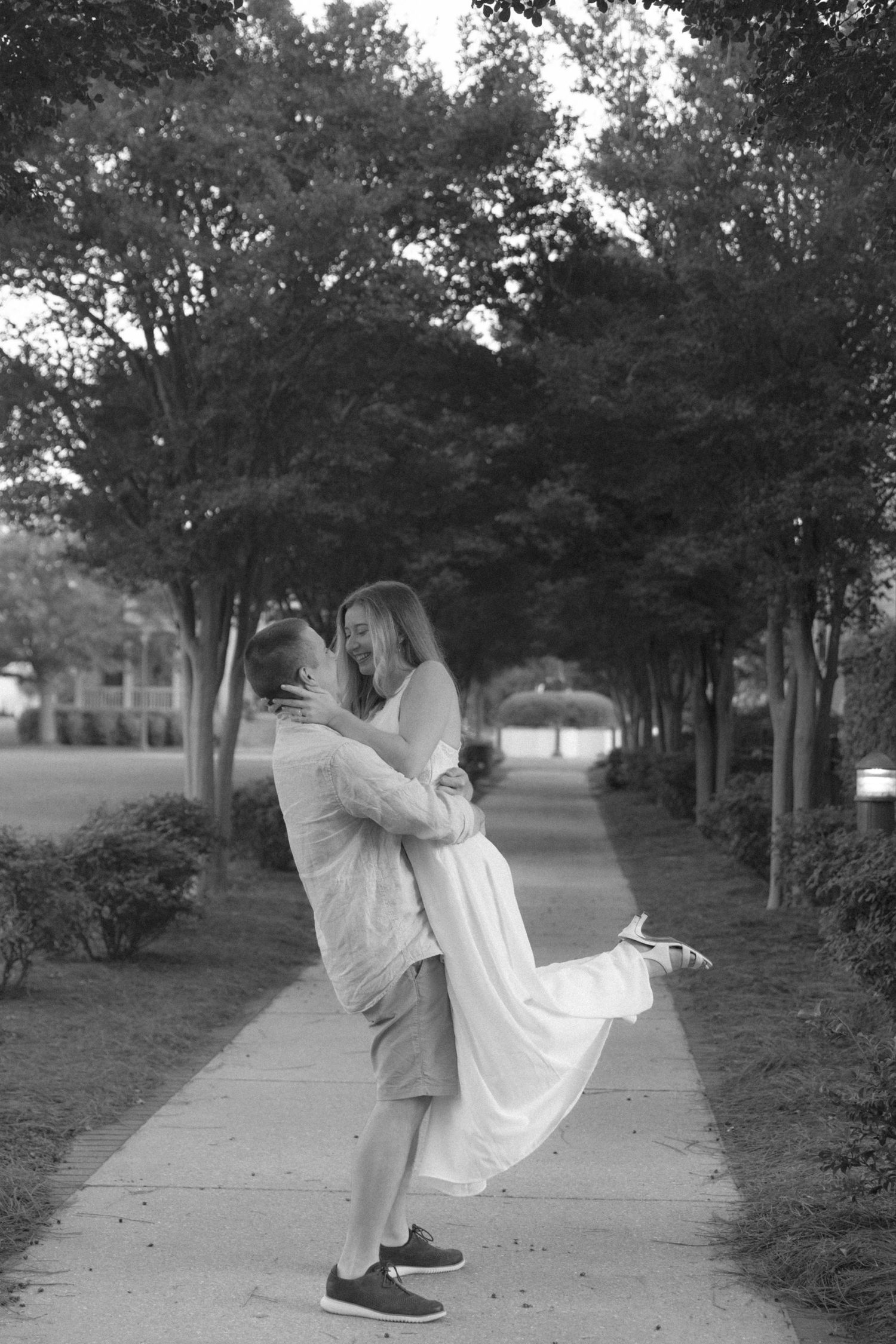 Black and white photo of joyful couple on a tree-lined path, man lifting woman in white dress.