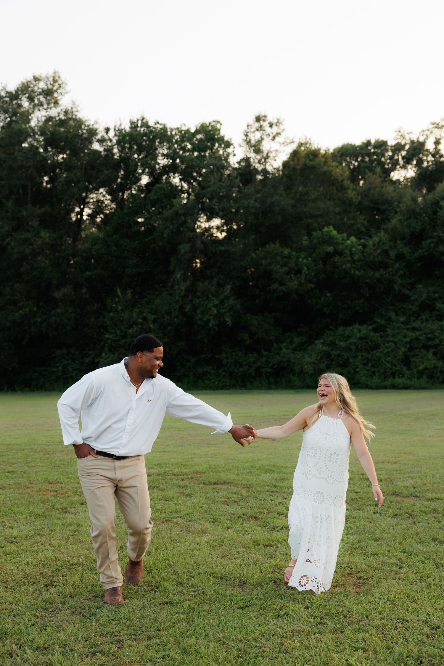 Laughing couple playfully holding hands while walking through a green field at dusk.