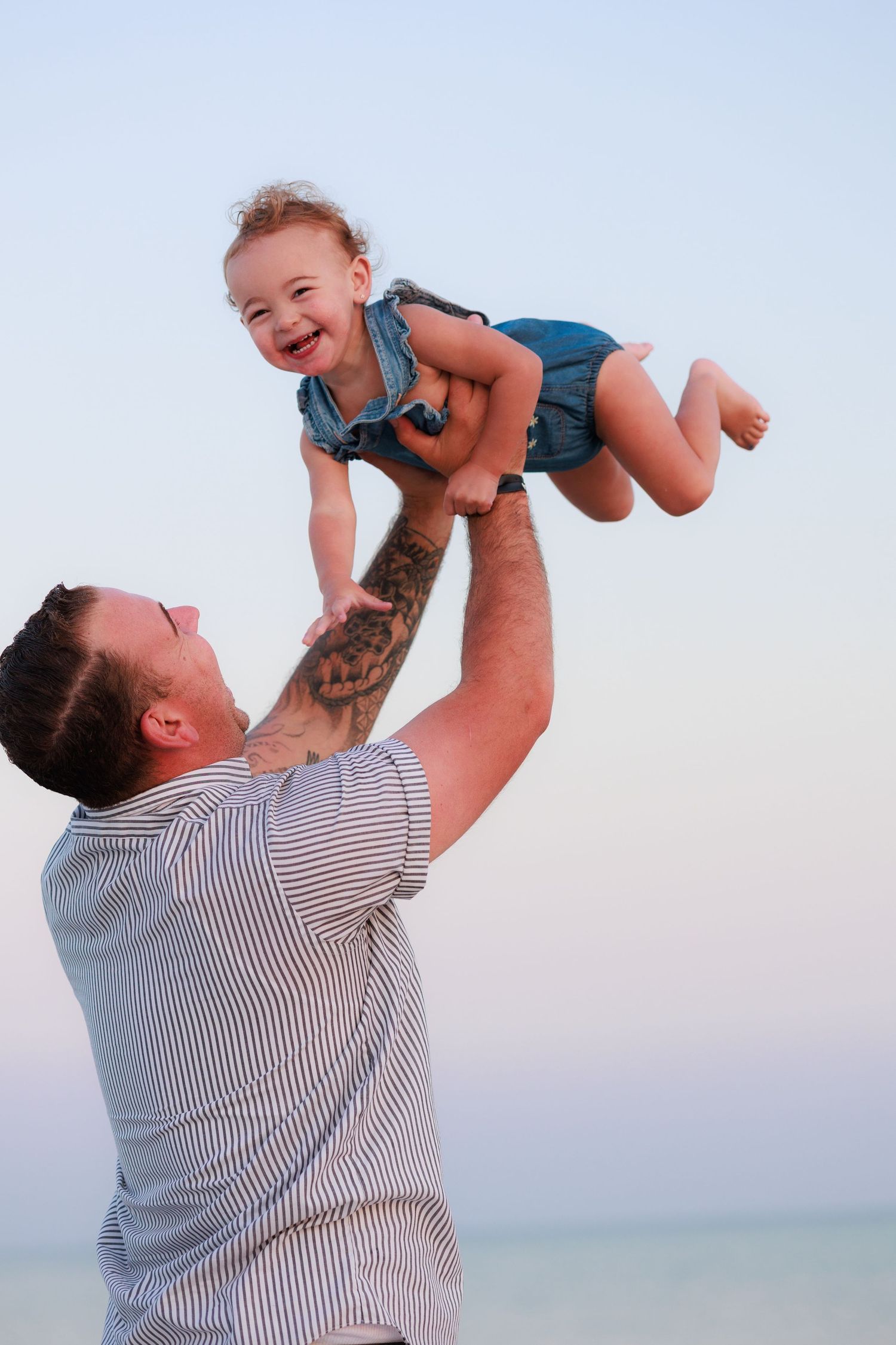 Father lifting laughing baby girl in the air at the beach during sunset, sharing a joyful moment.