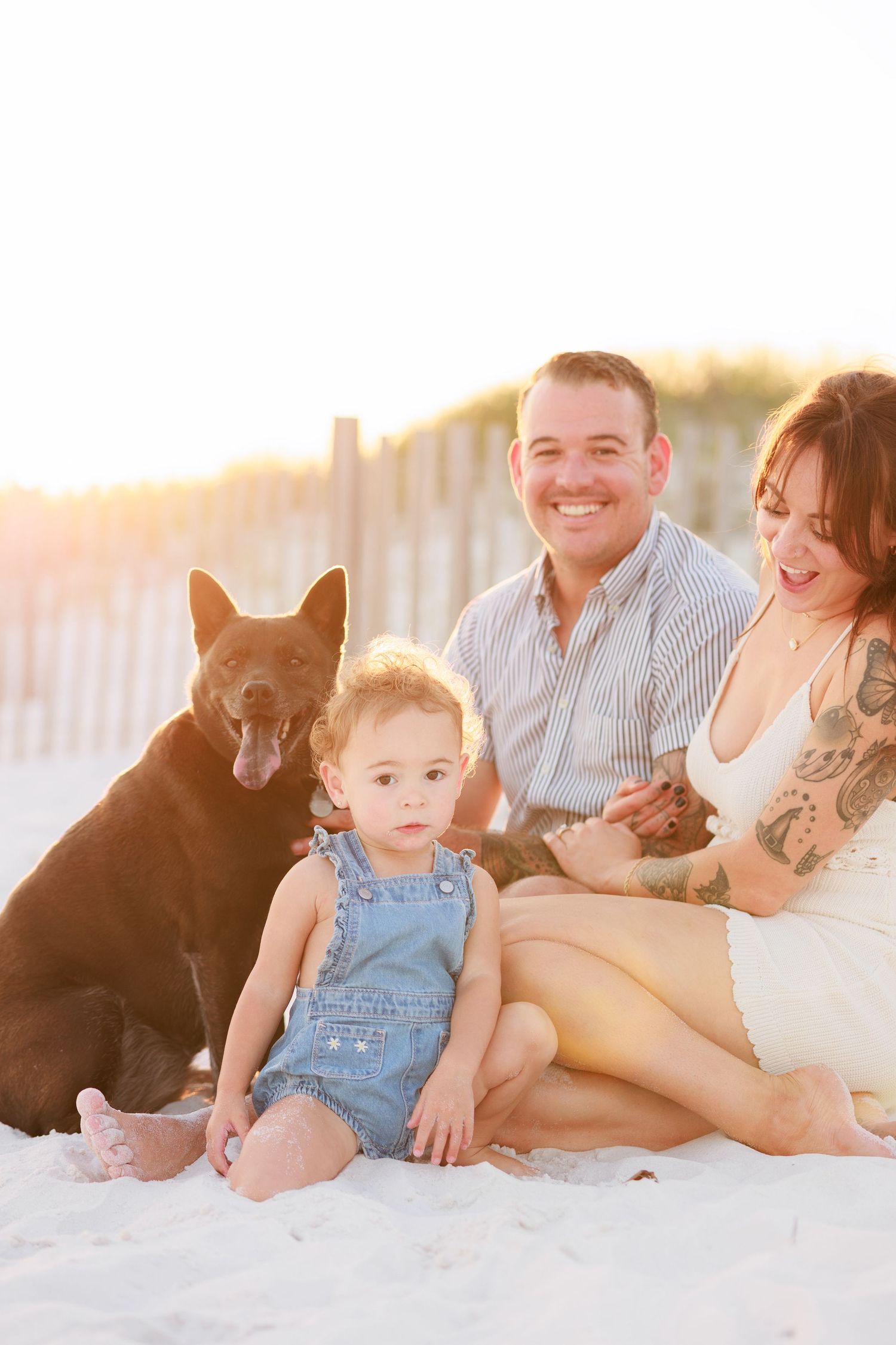 Family with toddler and brown dog posing on sandy beach at golden sunset hour.