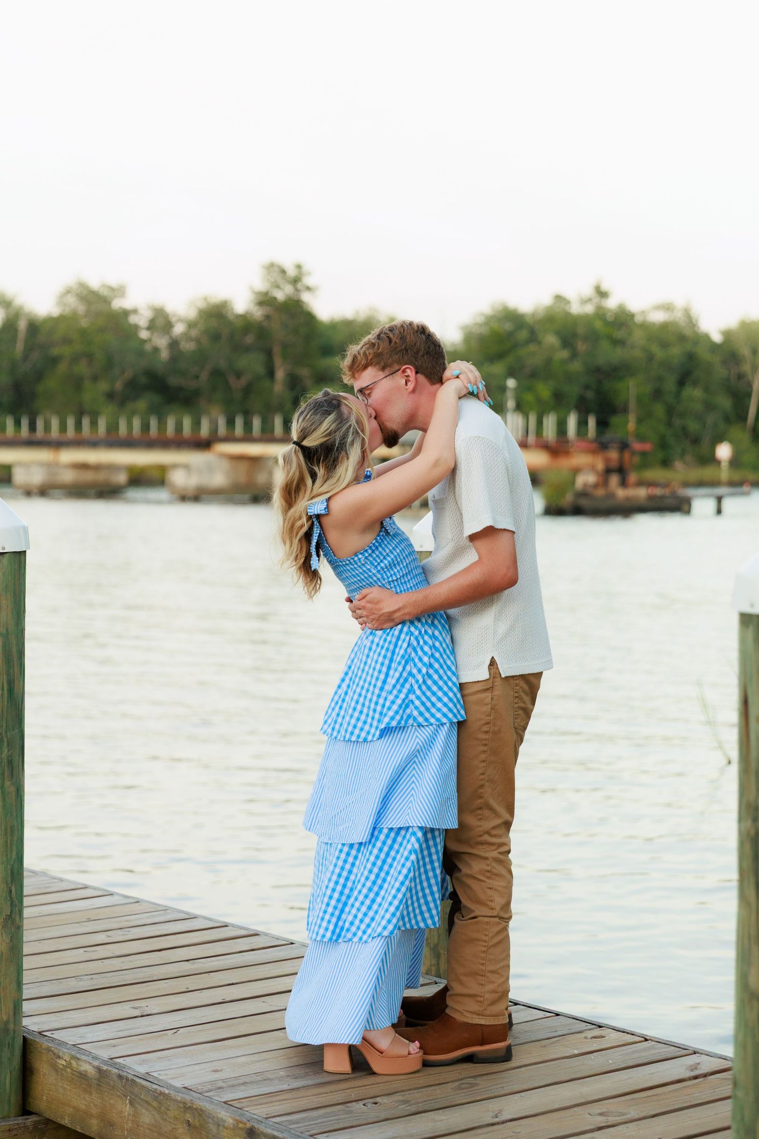 A couple shares a kiss on a wooden dock overlooking a serene waterway during their outdoor engagement session.