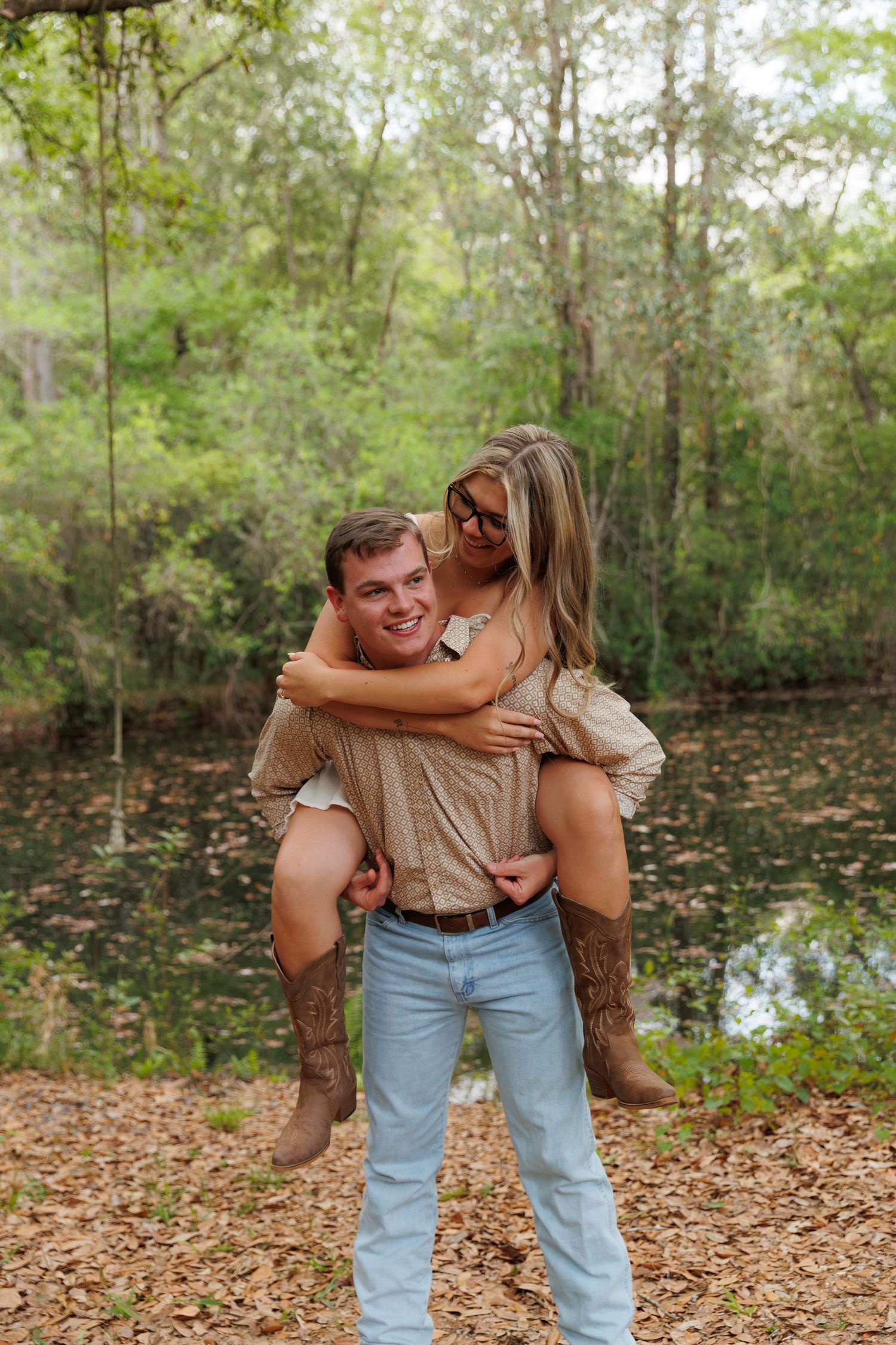 A smiling man gives a woman a piggyback ride outdoors near a pond surrounded by lush green trees.