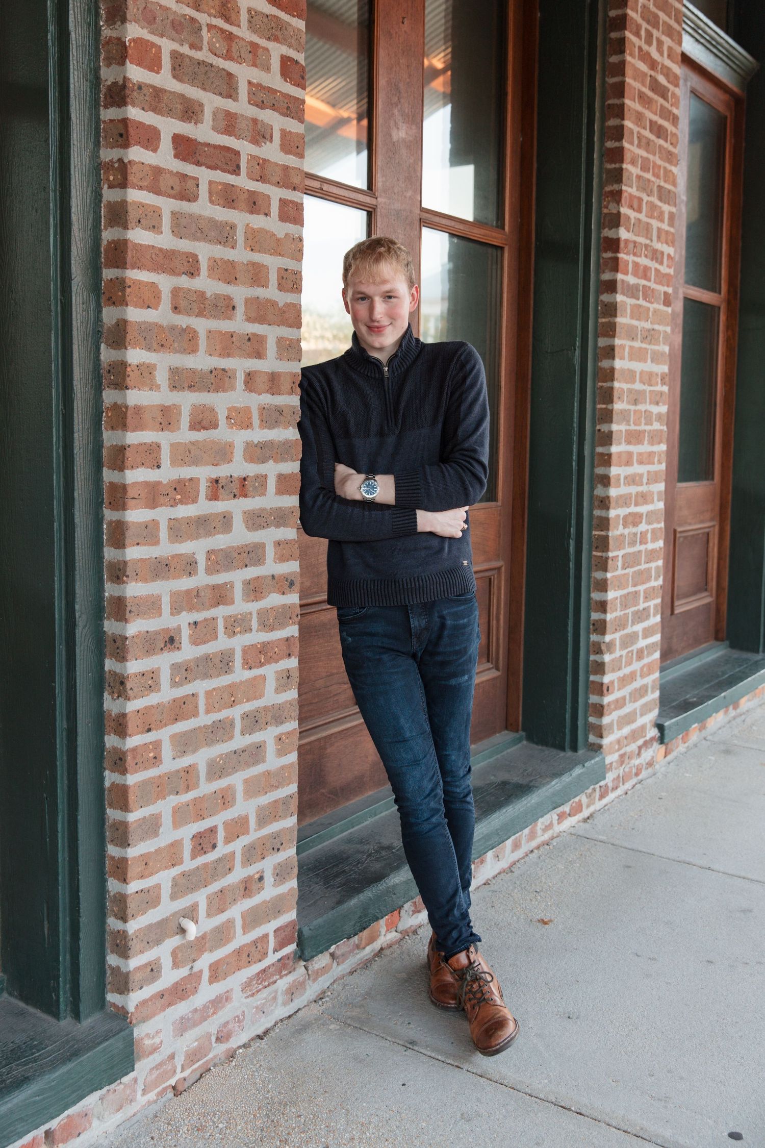 Young man in navy sweater and jeans leaning against brick building wall, wearing brown shoes and a watch.