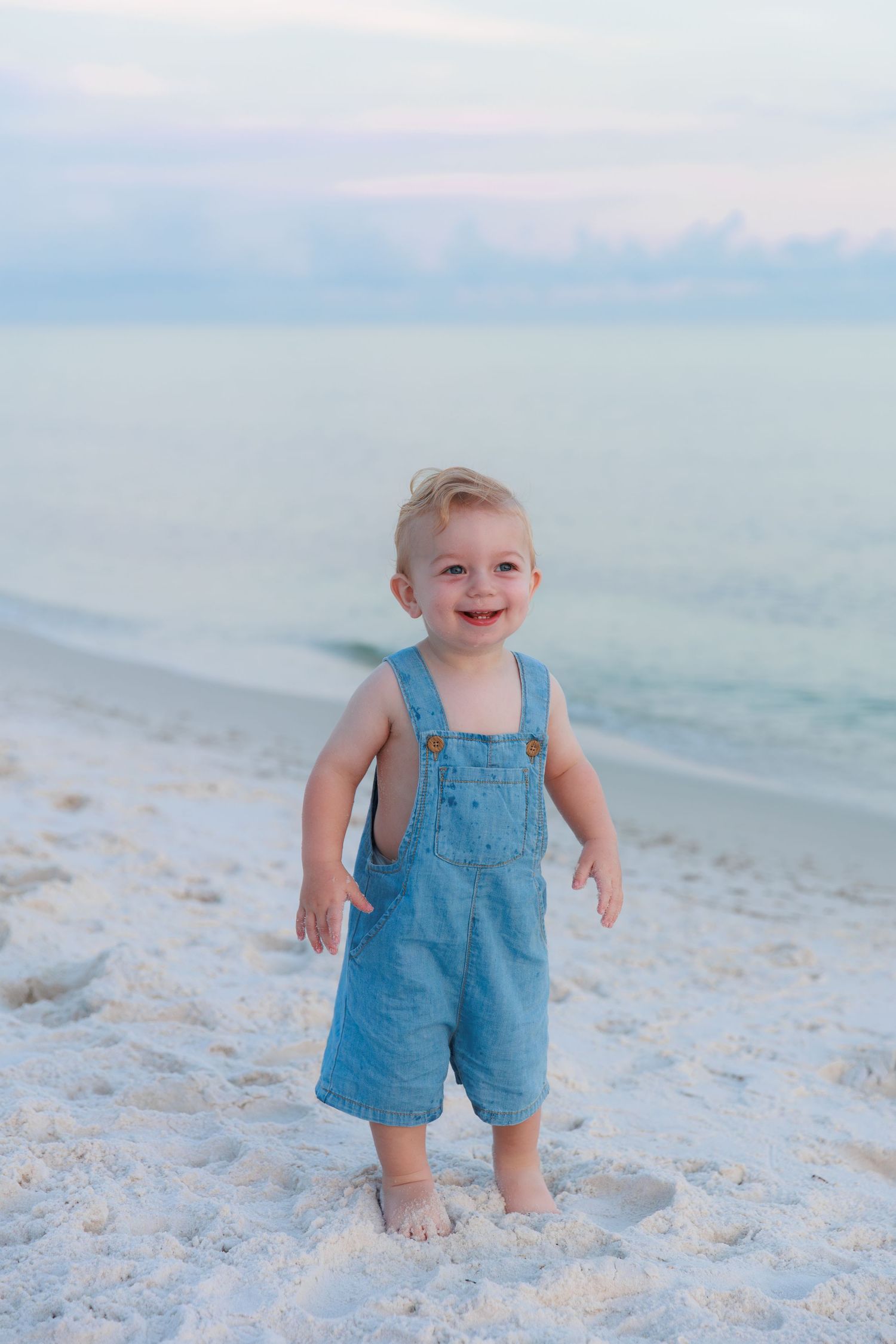 An adorable smiling toddler in denim overalls stands on a white sand beach with ocean waves behind him.