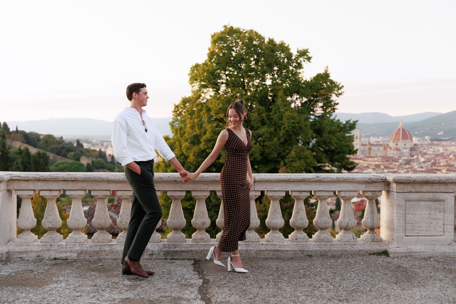 Couple holding hands on a stone terrace in Florence, Italy, with the Duomo and Tuscan hills in the background.