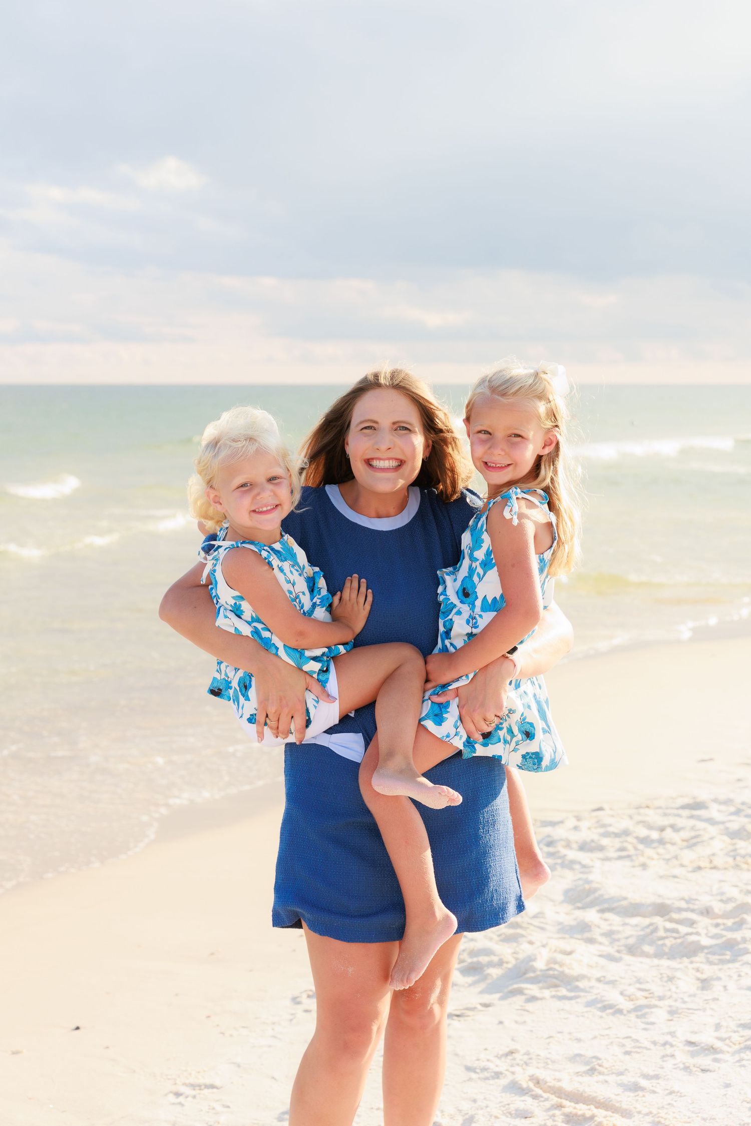 Smiling mother in blue dress holds two young daughters in matching floral dresses on a sunny beach.