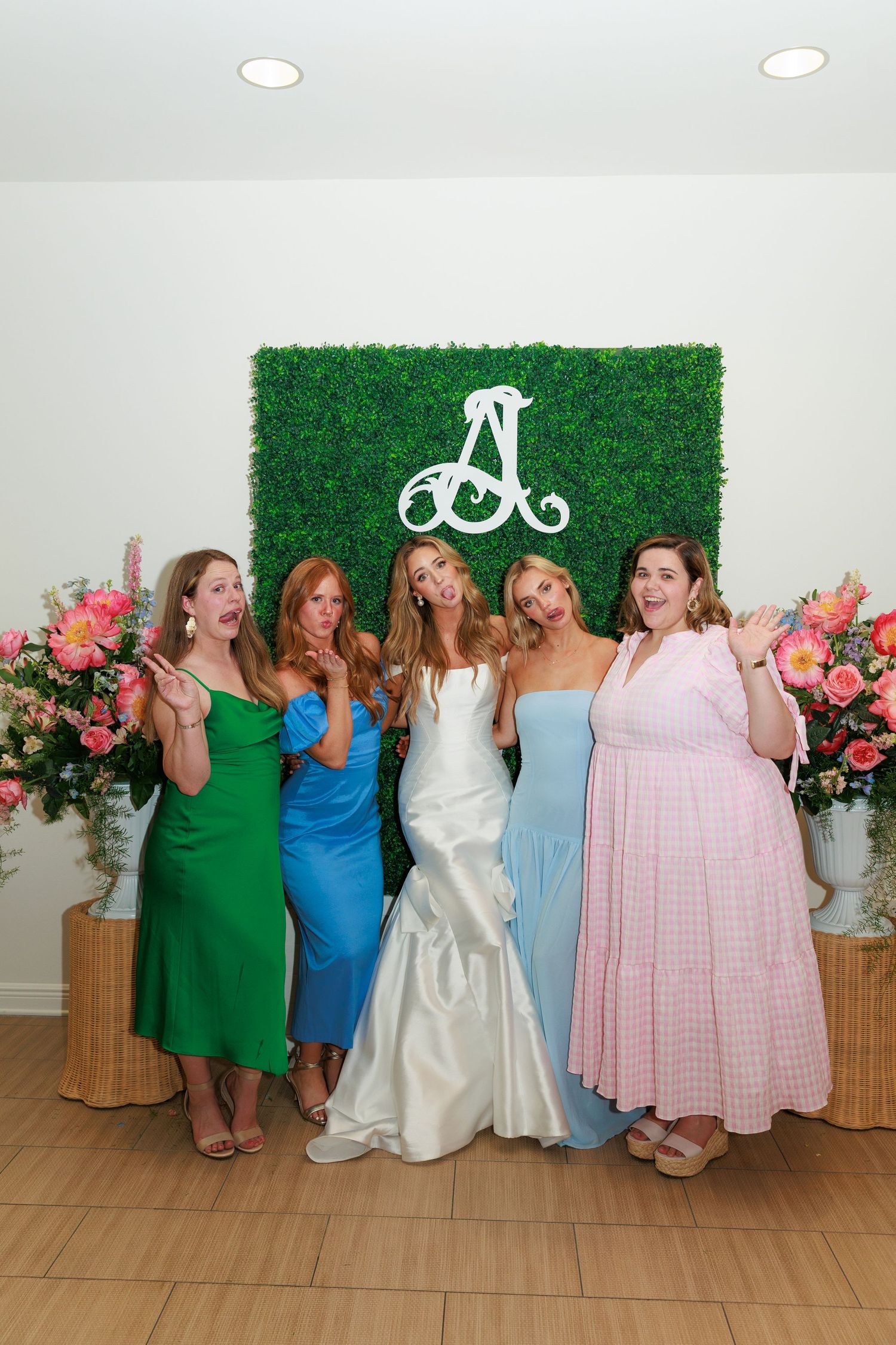 Bridesmaids in colorful dresses pose with bride in white gown against green backdrop with floral arrangements.