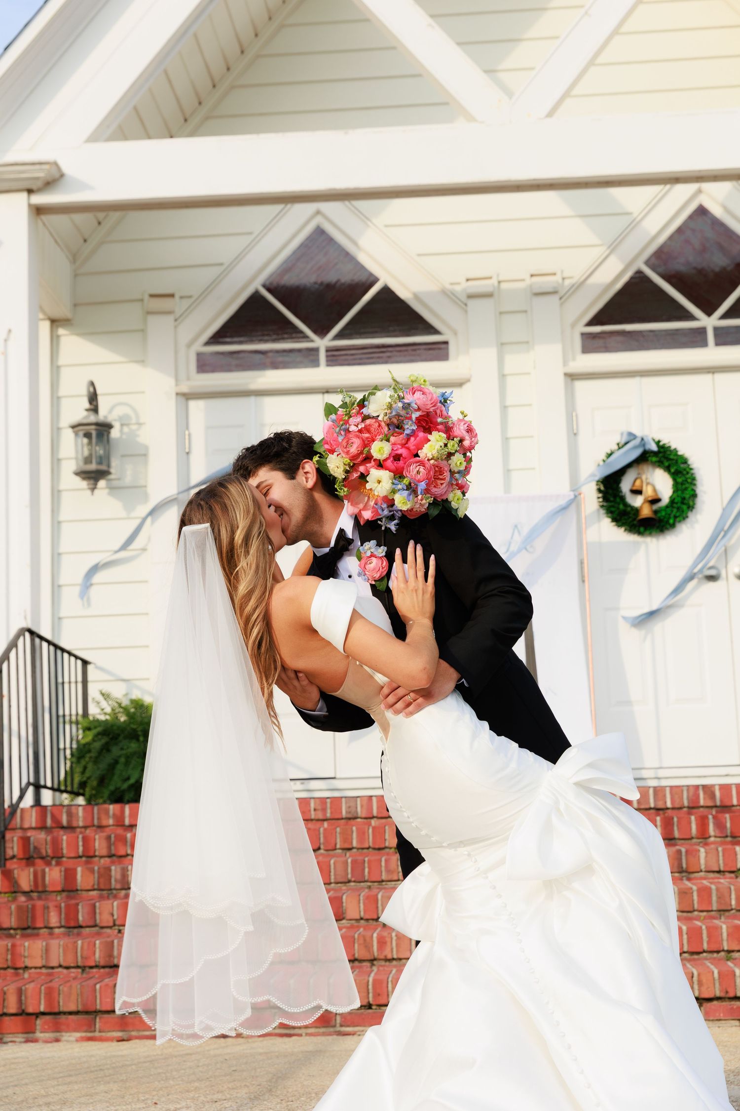 A romantic wedding photo showing the couple in a dip kiss outside a white chapel with a floral bouquet and green wreath.