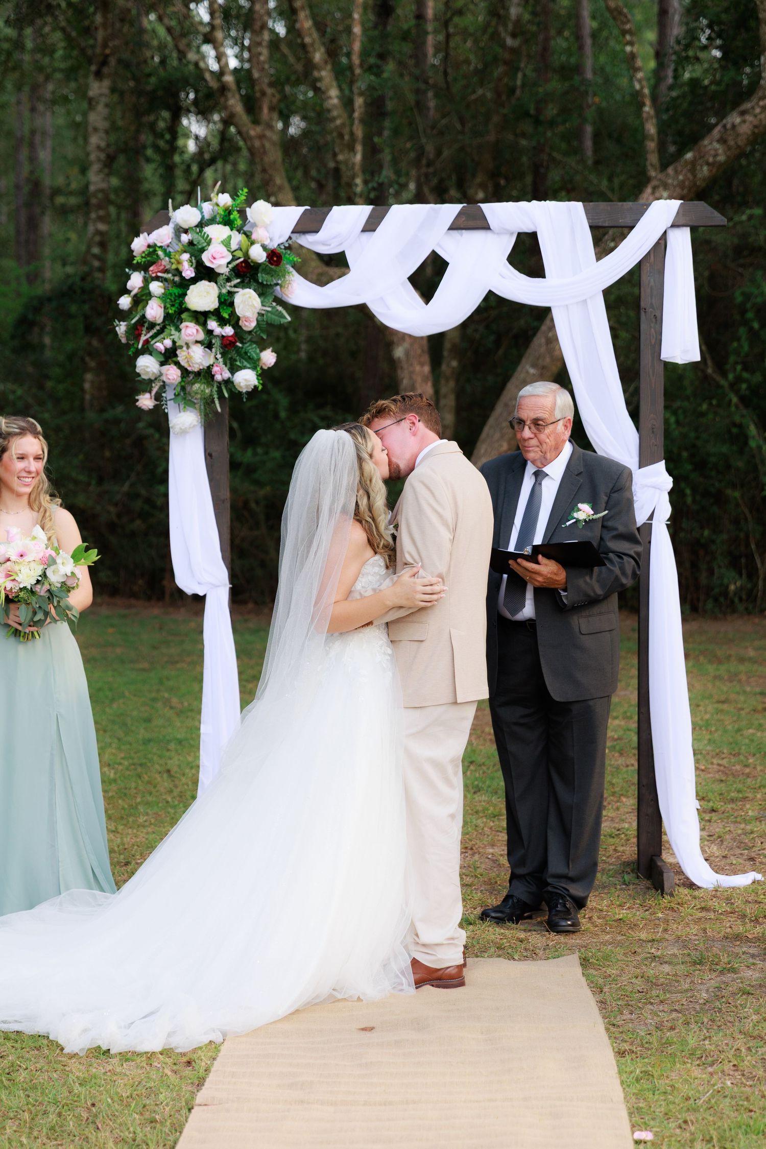 A romantic first kiss moment during an outdoor wedding ceremony beneath a white draped arch.