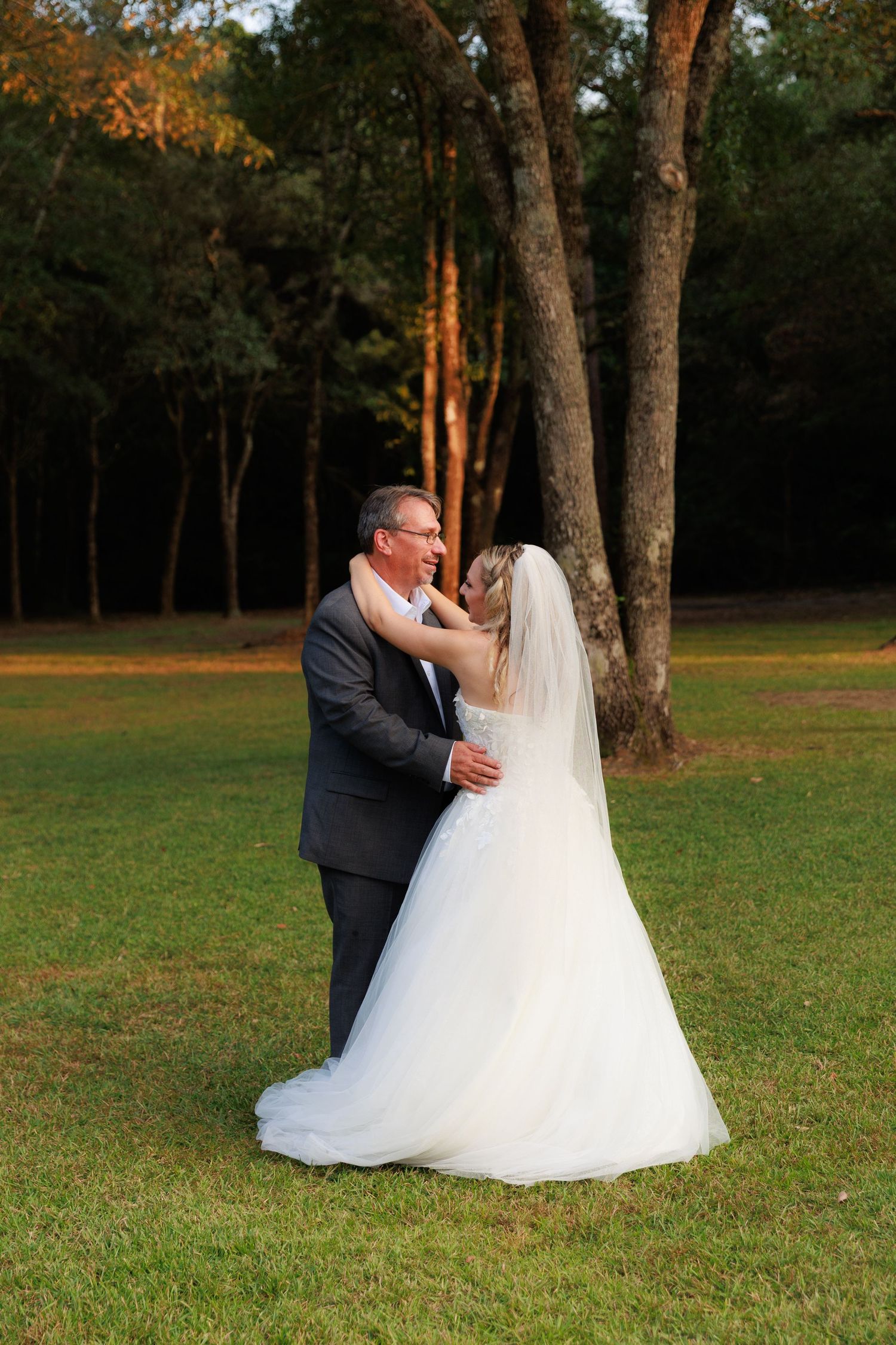 A couple in wedding attire dance together on a grassy lawn with trees in the background at sunset.