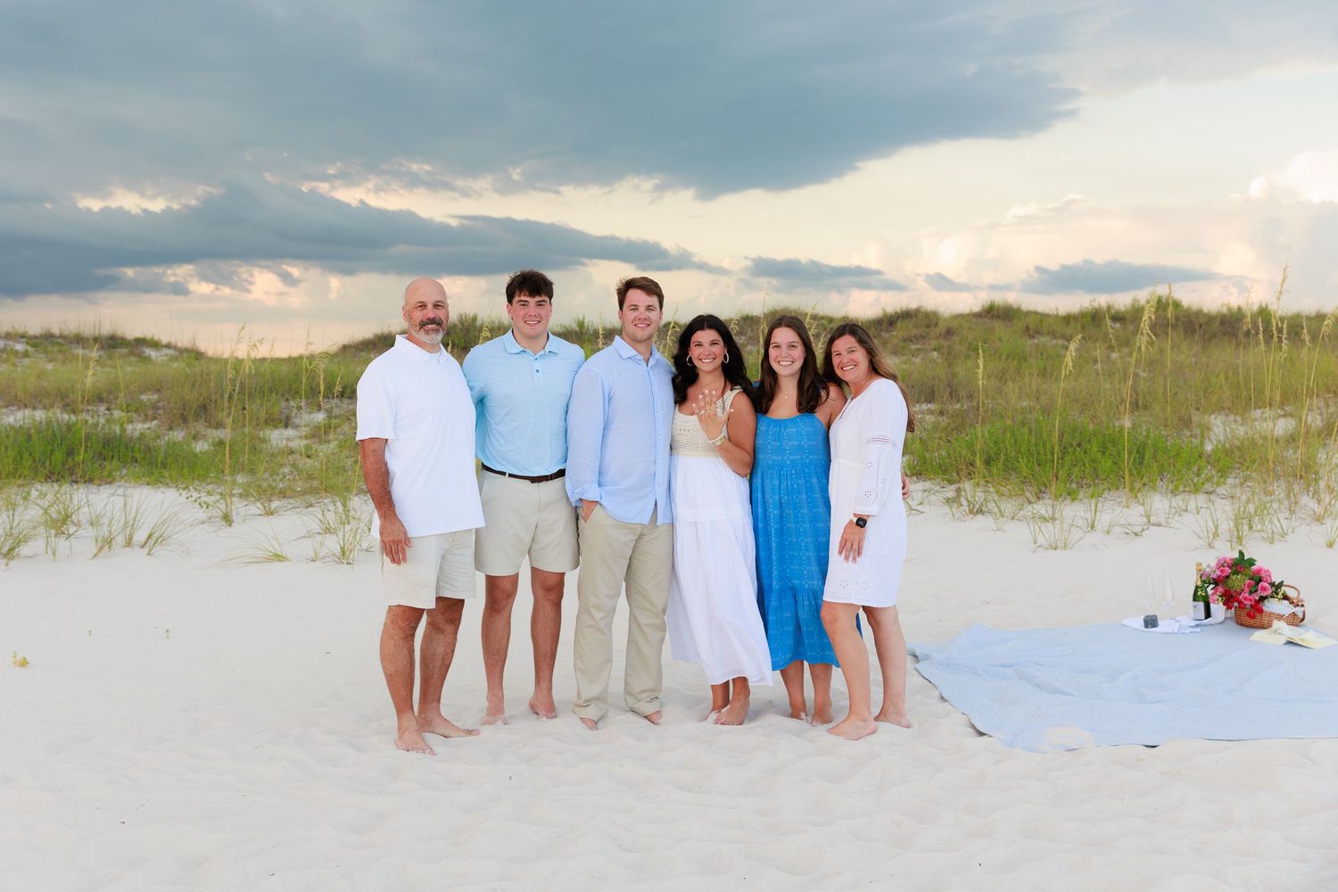 A group stands together on a white sandy beach with rolling dunes and blue sky in the background.