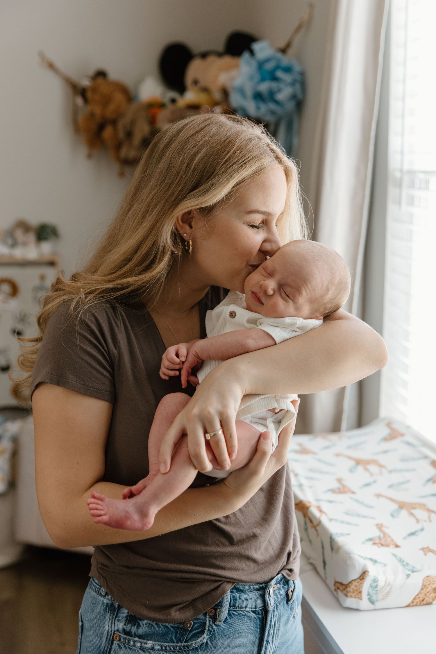A tender moment between parent and baby in a bright nursery room with stuffed animals visible in the background.