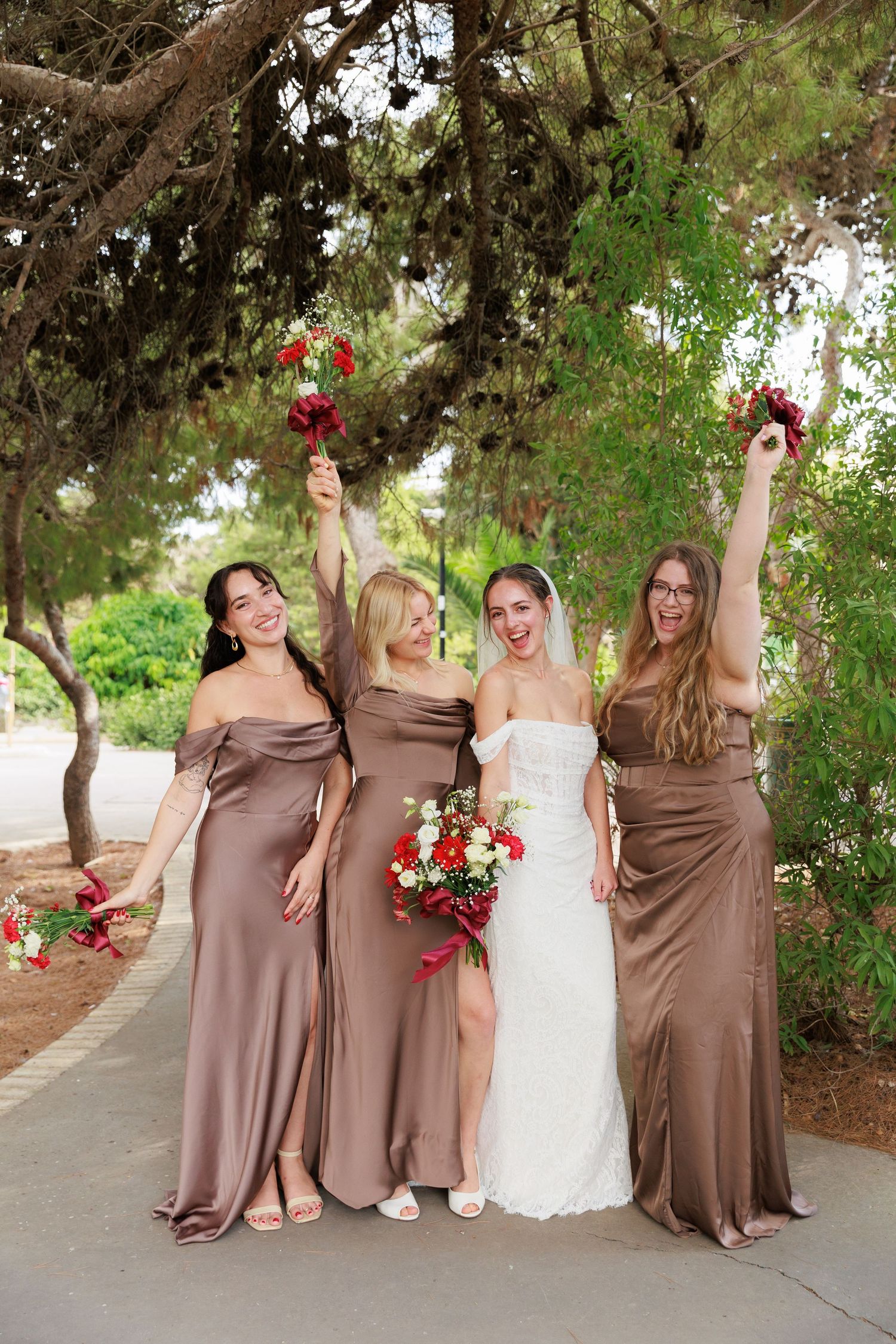 A bridal party in mocha-colored dresses and a white wedding gown pose joyfully with red floral bouquets on a garden path.