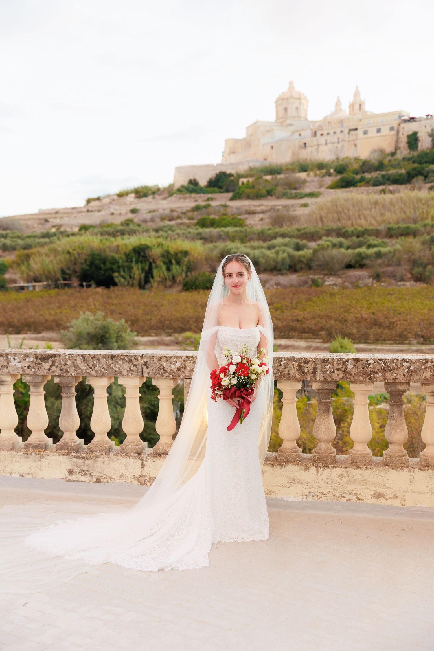 A bride in a white gown and veil holds a red bouquet while standing at a stone balustrade with a historic fortress in the background.