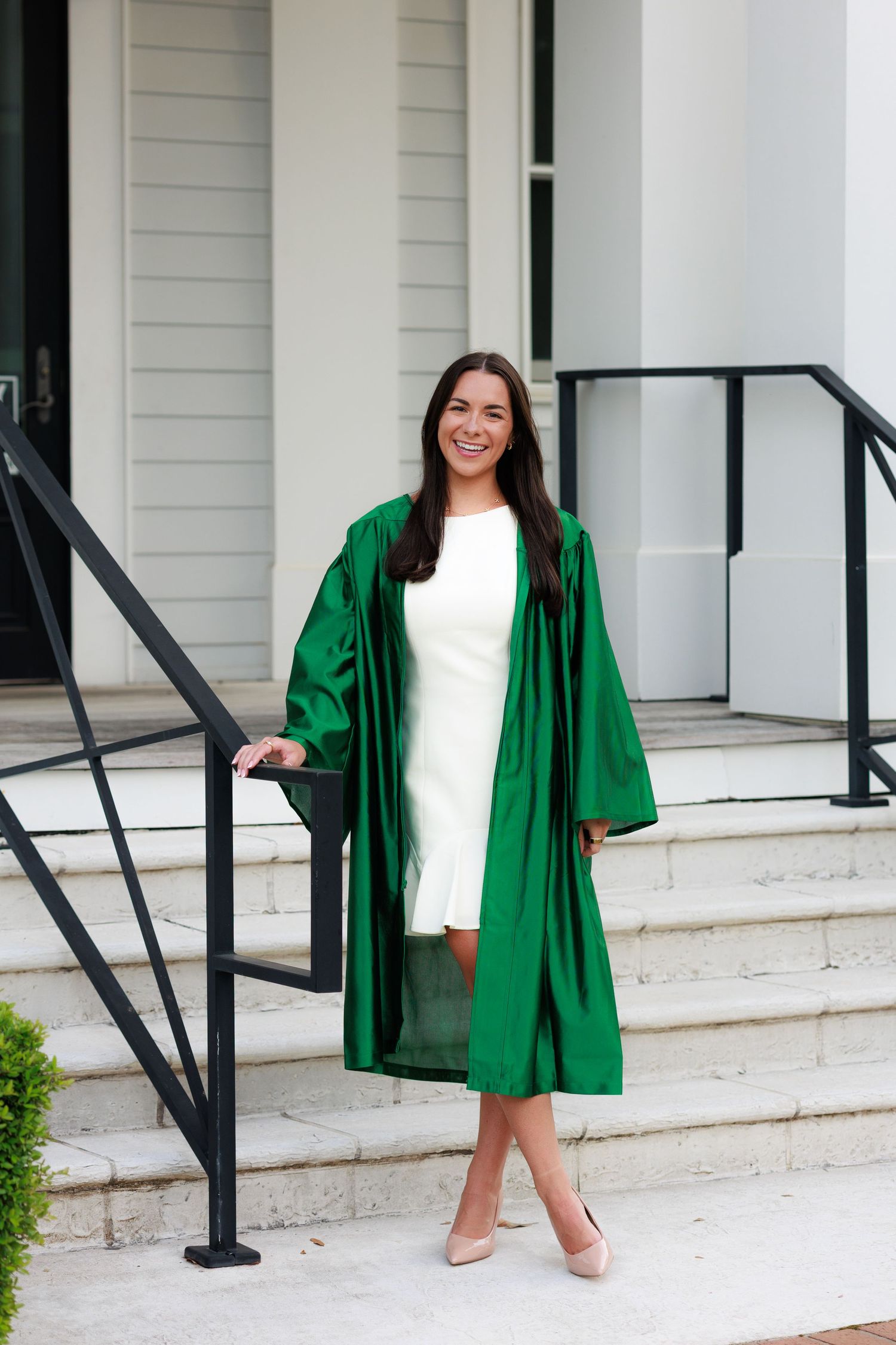 Smiling young woman in green graduation gown and white dress posing on white building steps.