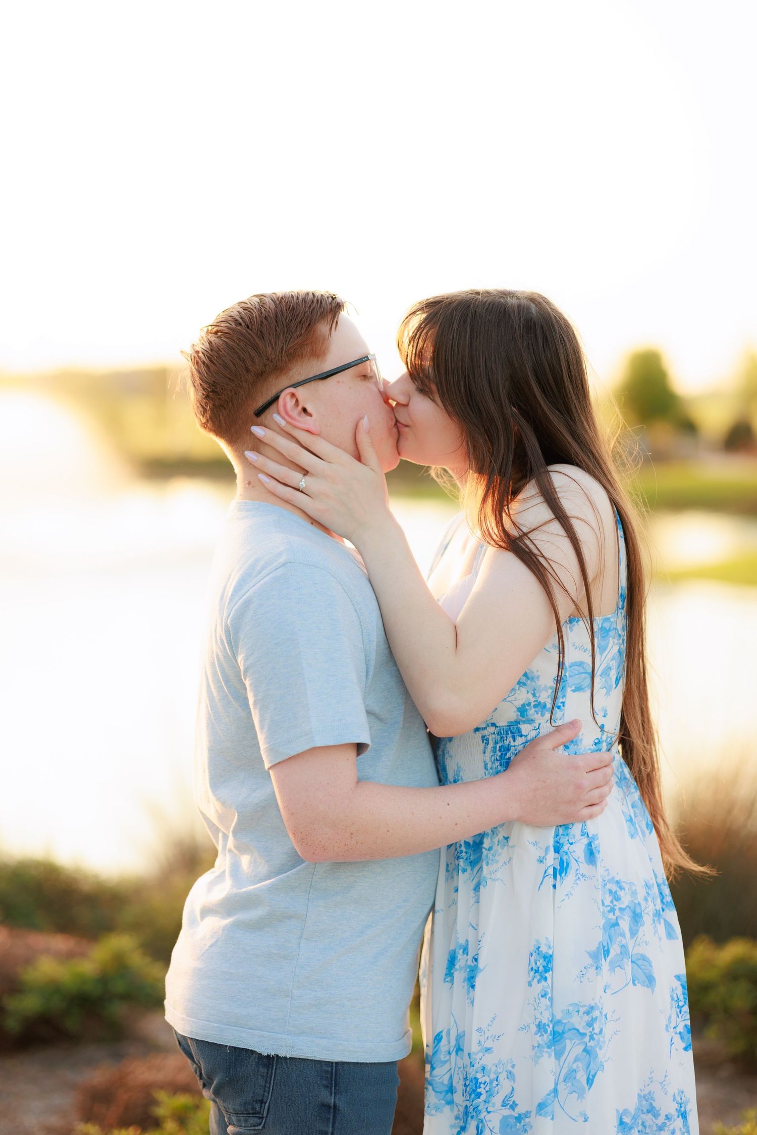 A young couple shares a romantic kiss outdoors near a lake at golden hour, woman in floral dress.